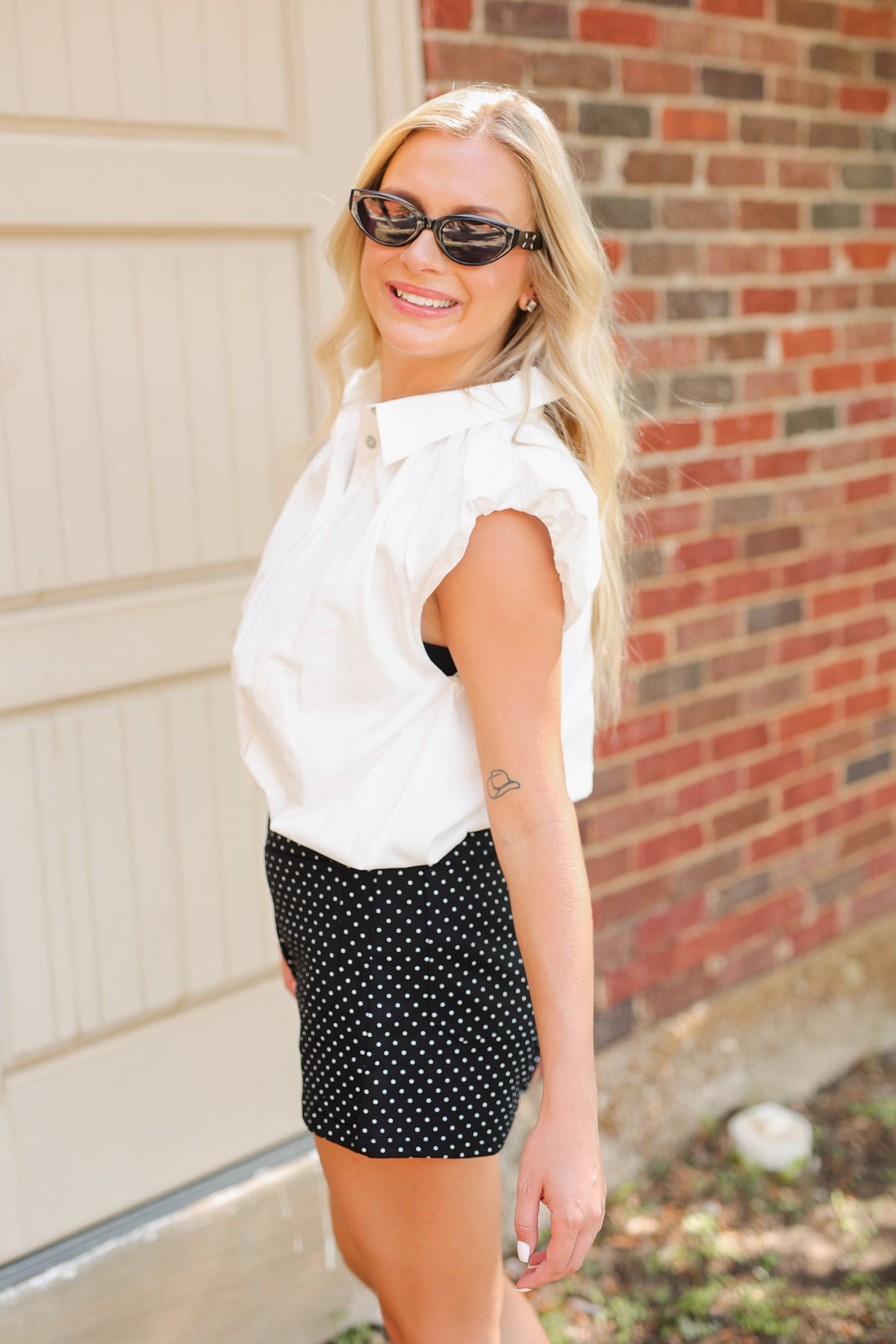 Blonde model wearing a sleeveless white top, black shorts with white dots and sunglasses standing outside.