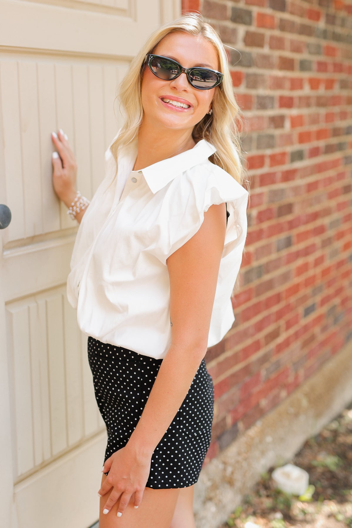 Blonde model wearing a sleeveless white top, black shorts with white dots and sunglasses standing outside.