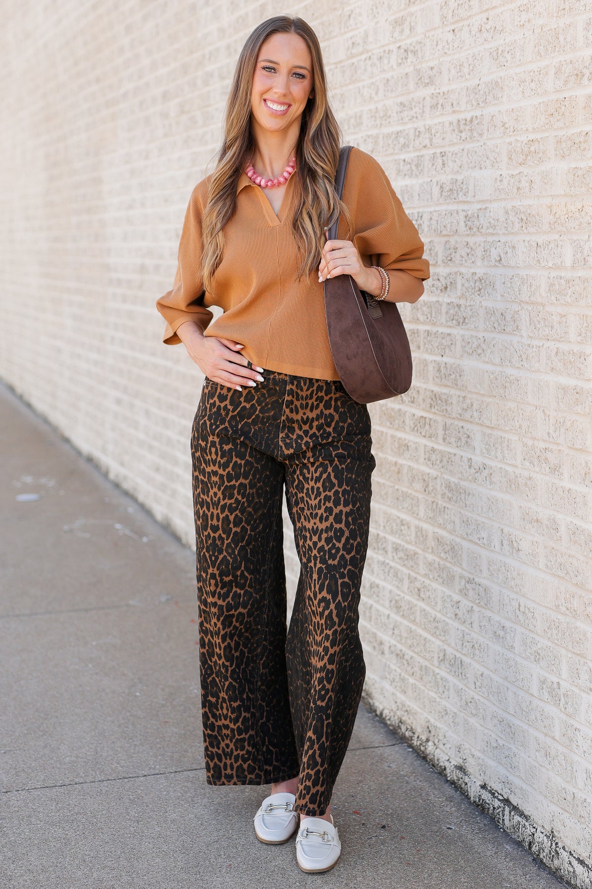 A woman stands against a light brick wall wearing a camel-colored blouse and wide-leg leopard-print pants, with a brown shoulder bag and white loafers.