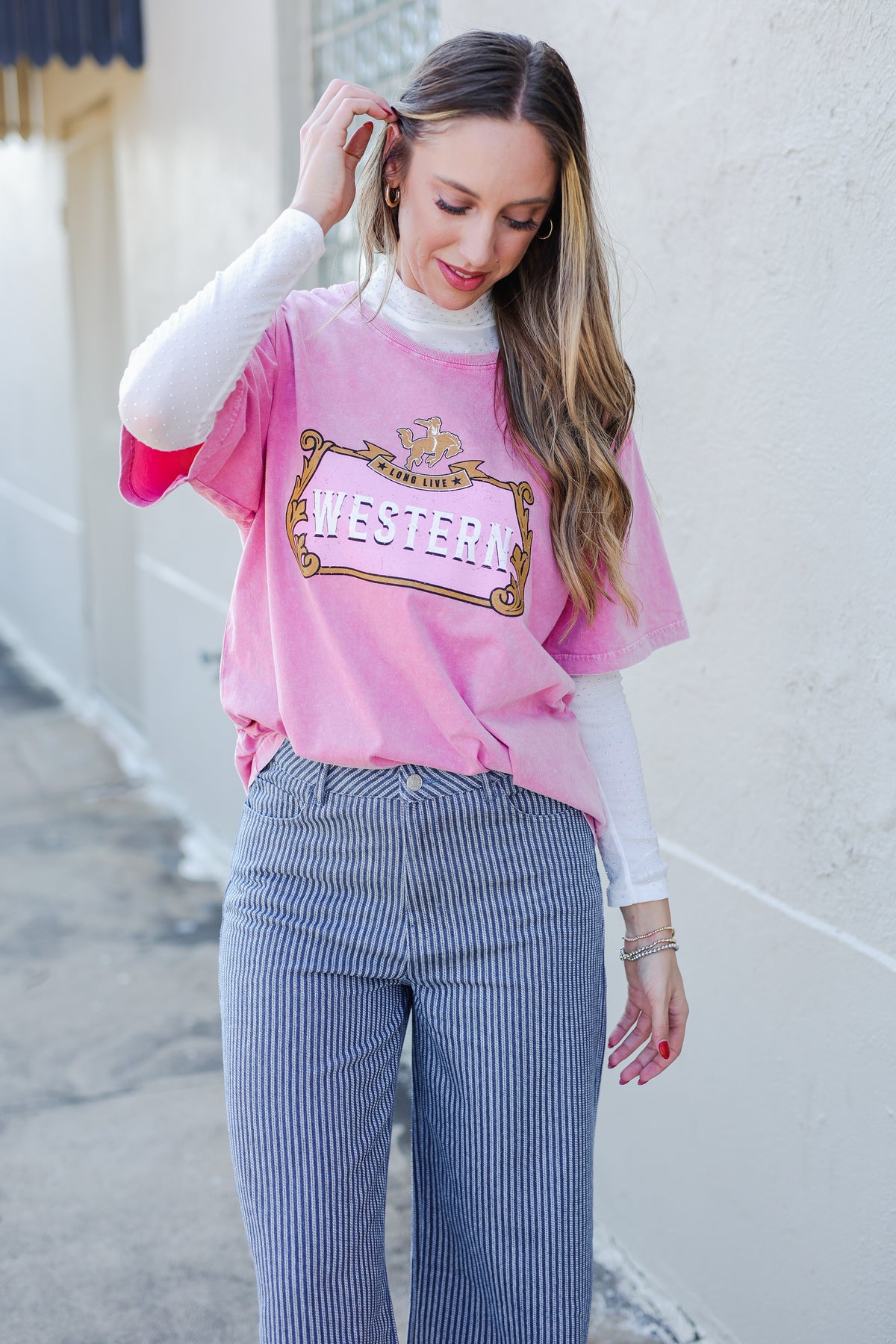 Woman wearing a pink oversized shirt with text and white long-sleeve underneath, standing against a light-colored wall.