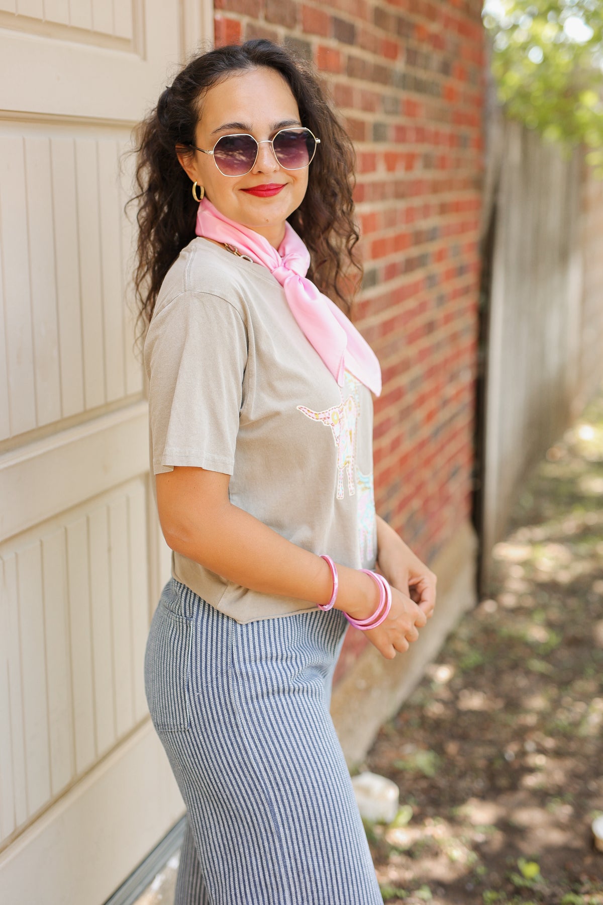 A woman wearing a light gray t-shirt with a pink neck scarf, blue and white striped skirt, and sunglasses, posing outdoors next to a brick wall and beige garage door.