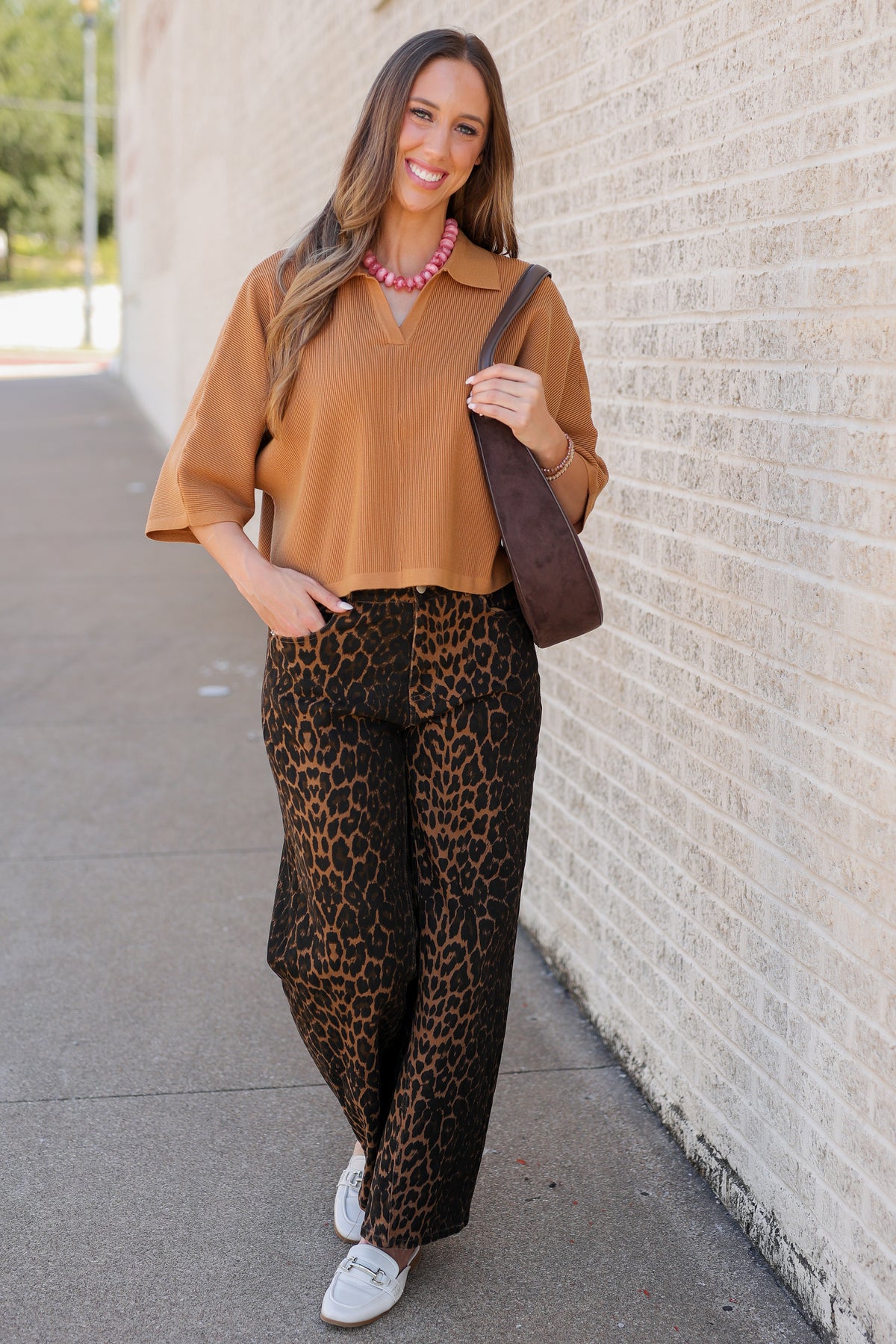 A woman stands on a sidewalk next to a light brick wall, wearing a tan short-sleeve collared top and brown leopard-print pants, carrying a dark brown shoulder bag.