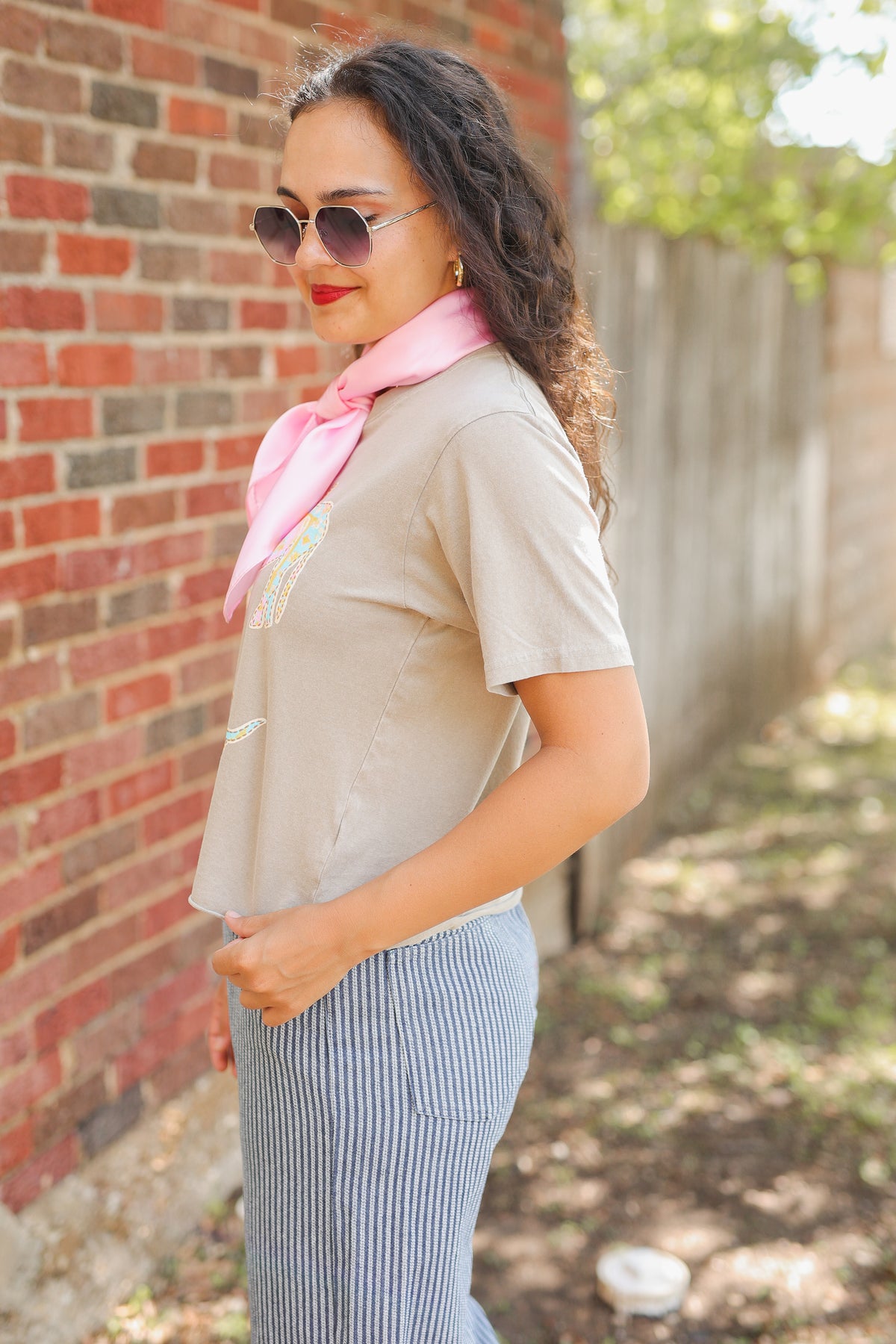 A woman wearing a beige short-sleeve top, a pink neck scarf, and blue-and-white striped pants, standing outdoors next to a brick wall and wooden fence.