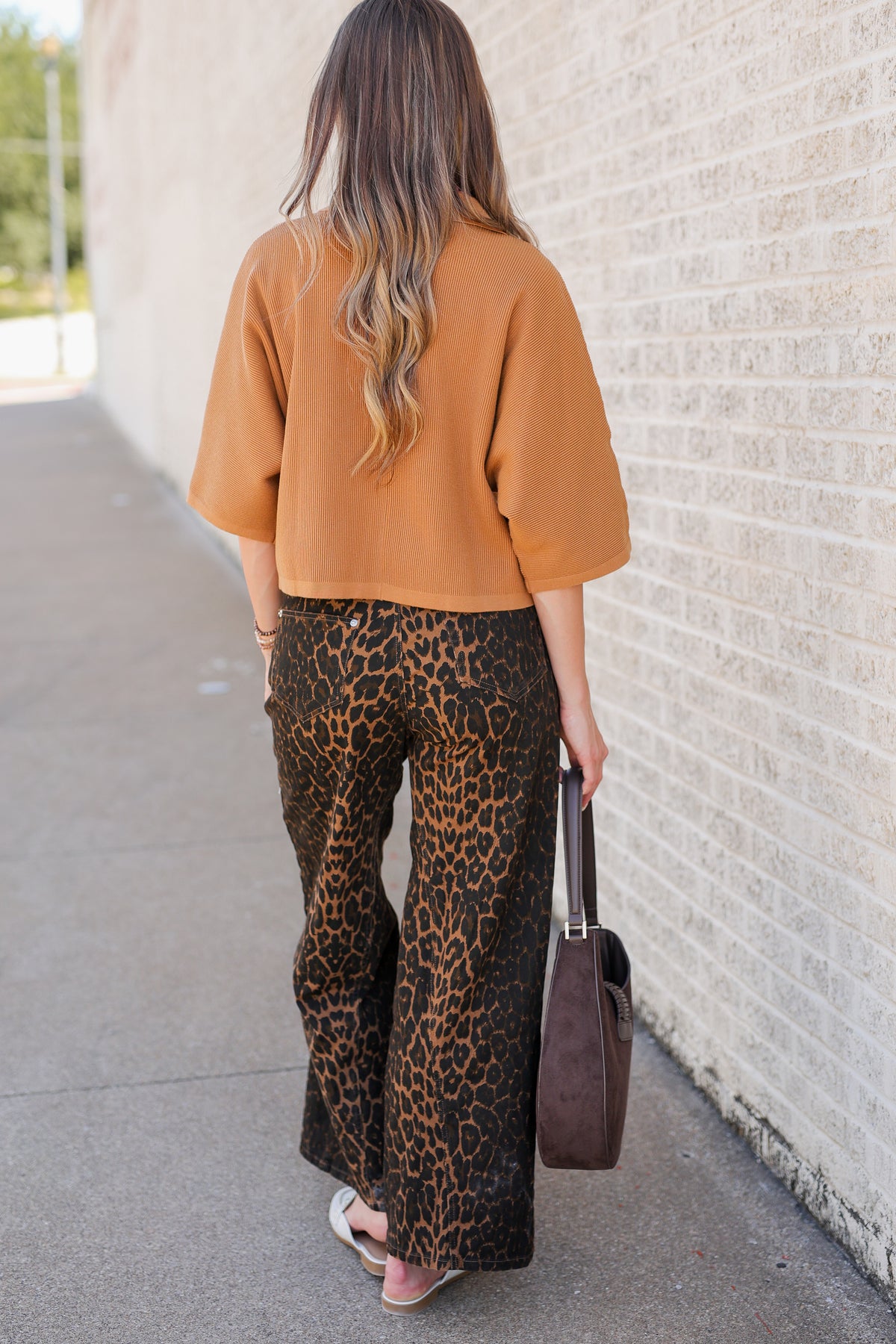 A woman viewed from behind wearing a mustard yellow ribbed knit top and brown leopard-print wide-leg pants, standing on a sidewalk next to a light brick wall and holding a dark brown handbag.