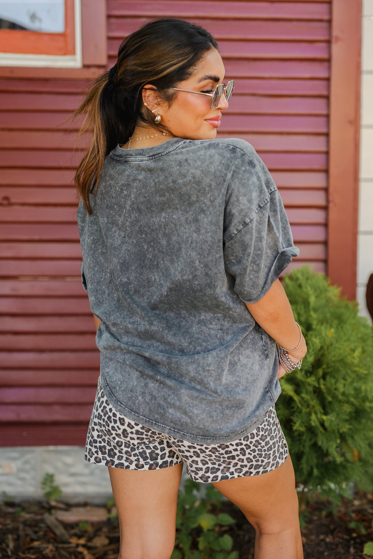 Woman wearing a gray tie-dye shirt and leopard print shorts standing in front of a red building.