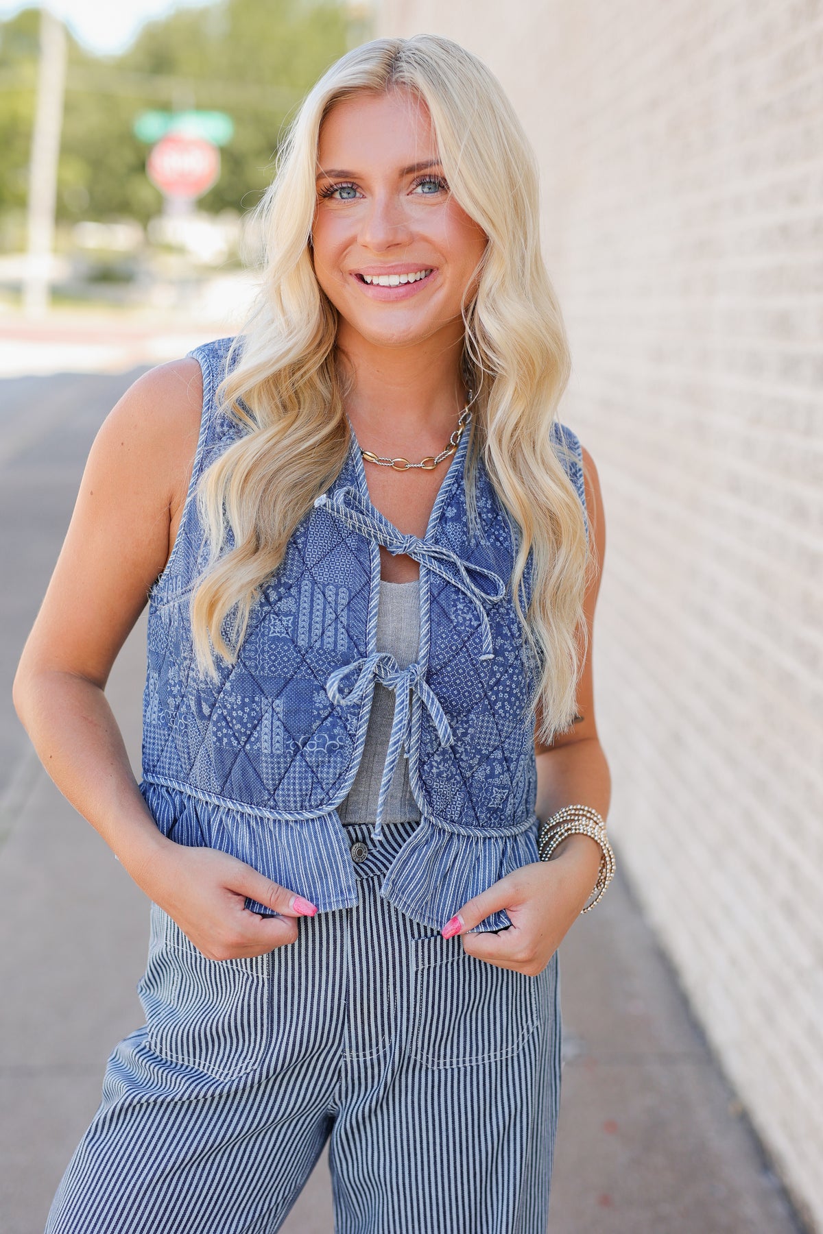 A smiling blonde woman wearing a sleeveless blue patterned vest over a light top and blue-and-white pinstripe pants, standing outdoors against a light brick wall.