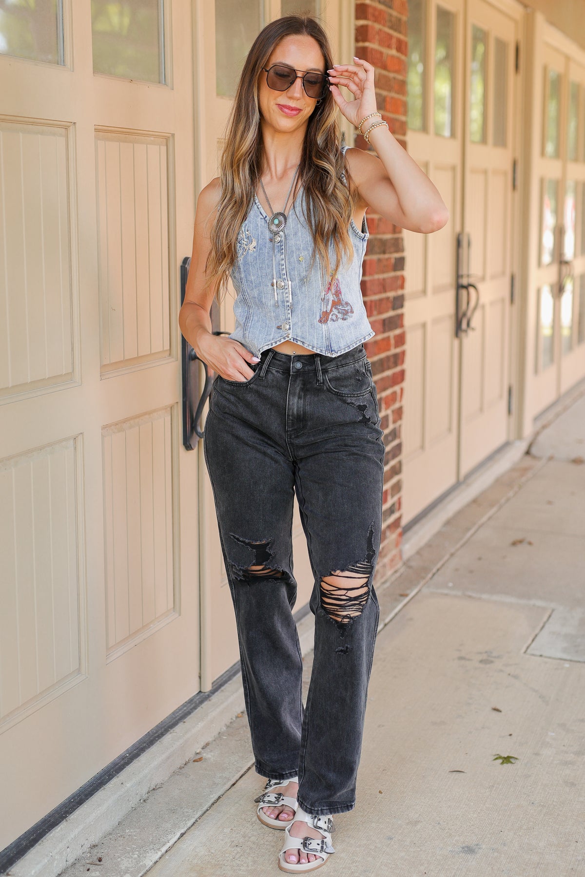 A woman stands on a sidewalk in front of beige garage doors and a brick wall, wearing a sleeveless light blue denim vest with floral embroidery, dark gray distressed jeans with ripped knees, and white slide sandals.