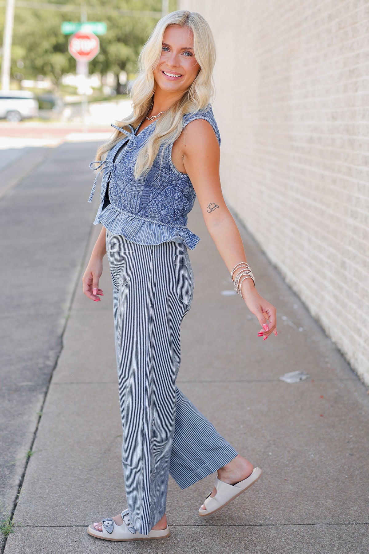 A woman modeling a two-piece outfit consisting of a light blue denim ruffle-trim top and matching blue-and-white vertically striped pants, walking on a sidewalk next to a brick wall.