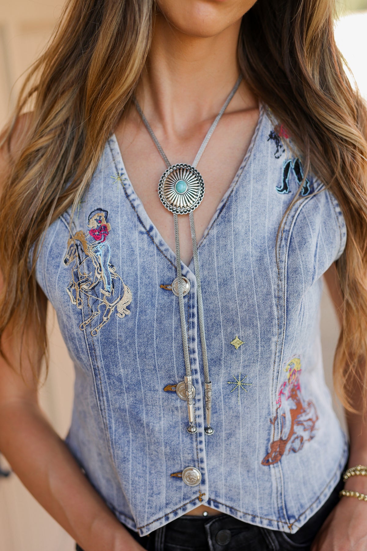 A close-up of a woman wearing a light blue denim vest with western embroidery, silver-toned buttons, and a long silver-toned necklace with a round pendant.