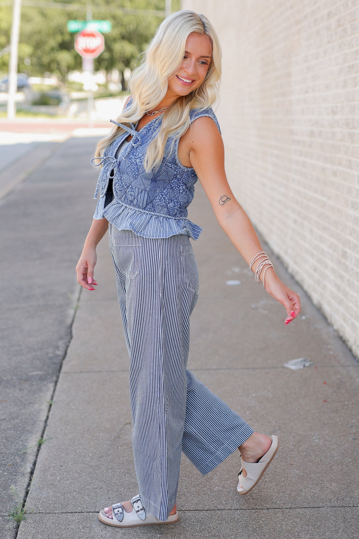 A woman posing on an outdoor sidewalk wearing a light blue denim vest and matching blue-and-white striped pants.