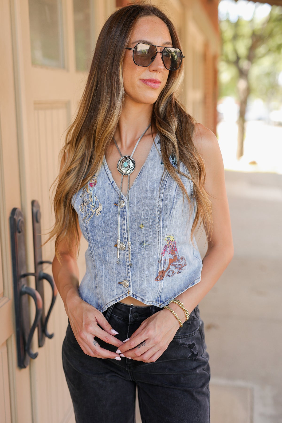 A woman wearing a sleeveless light-wash denim vest with colorful embroidered details, paired with dark jeans and sunglasses, standing outdoors.