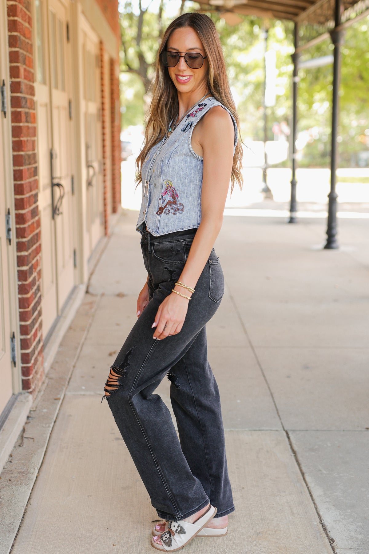A woman modeling a sleeveless light blue denim vest with colorful floral embroidery over a white top, paired with dark gray distressed jeans and white slide sandals, standing on a sidewalk next to a brick building.