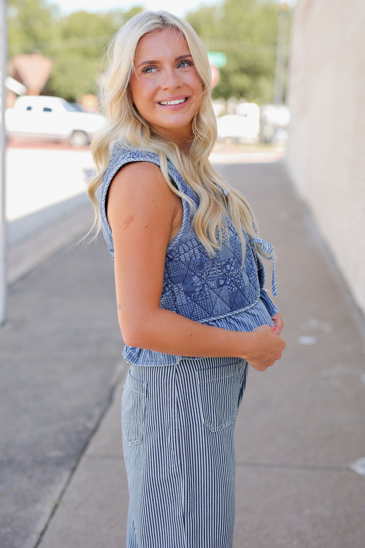 A smiling blonde woman wearing a sleeveless blue patterned top and matching blue-and-white striped pants, standing outdoors on a sidewalk with a blurred street background.