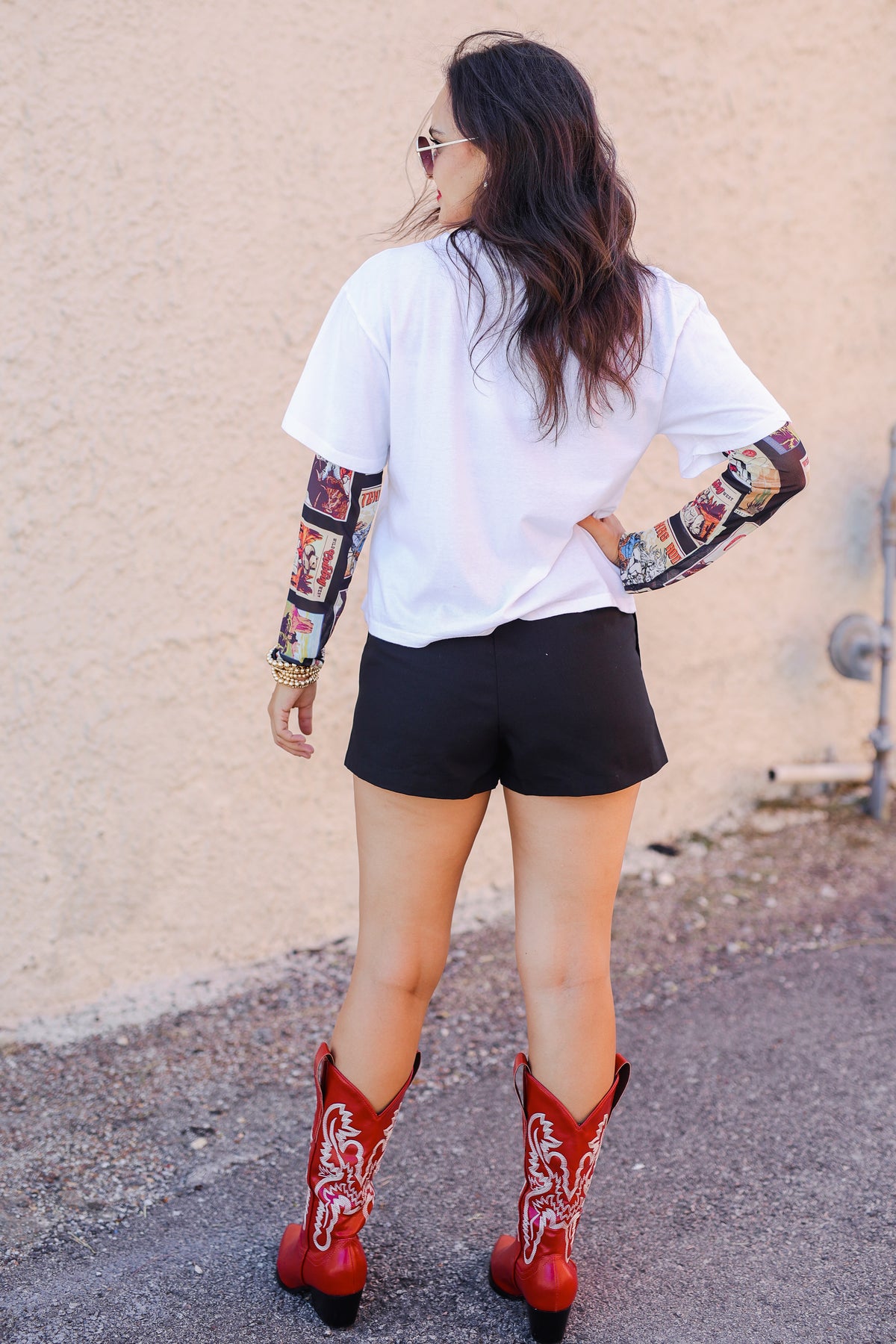 Person wearing a white t-shirt with colorful sleeves, black shorts, and red cowboy boots against a beige wall.