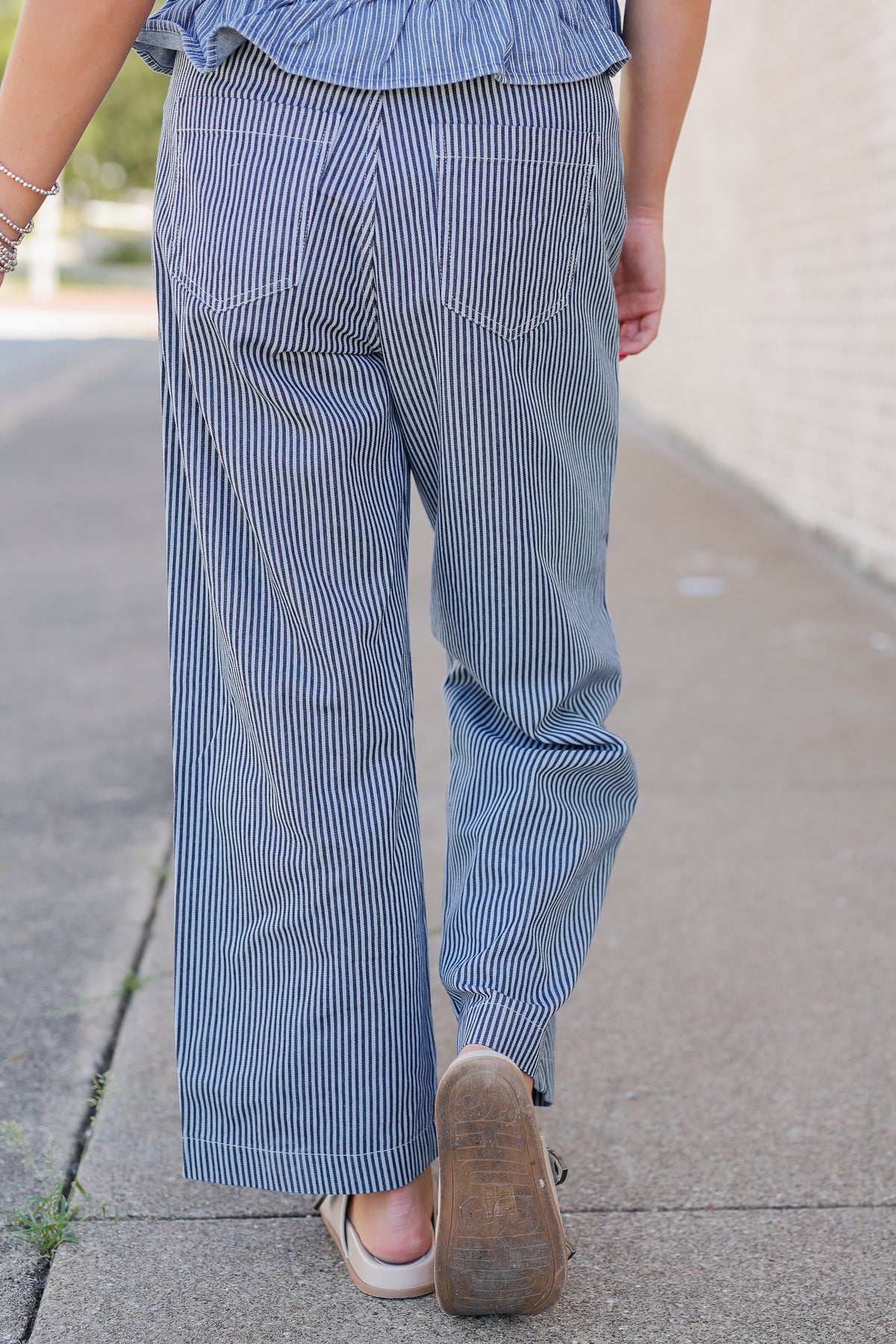 A person viewed from behind wearing blue and white vertically striped wide-leg pants, a matching striped top, and brown leather sandals on a concrete sidewalk.
