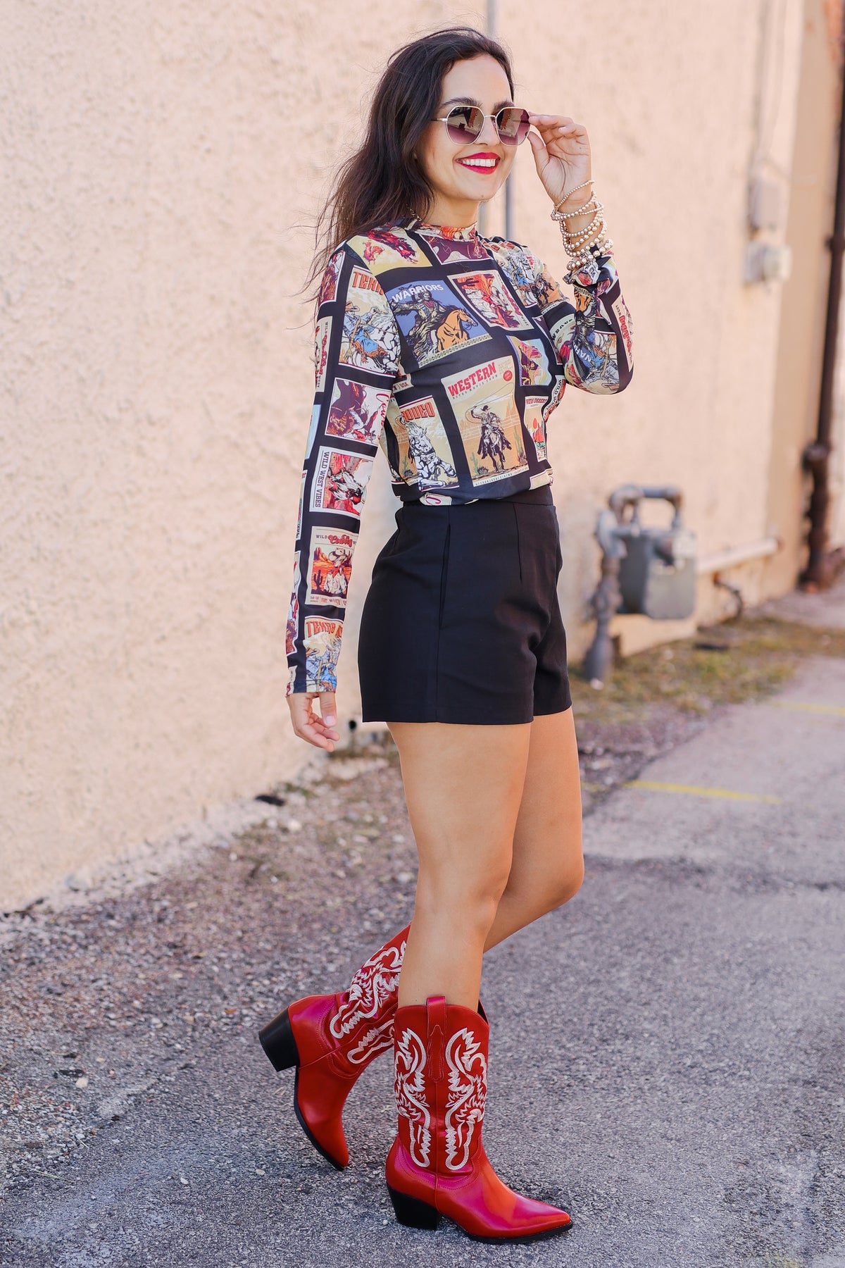 Woman wearing a colorful patterned blouse and red cowboy boots standing against a beige wall.