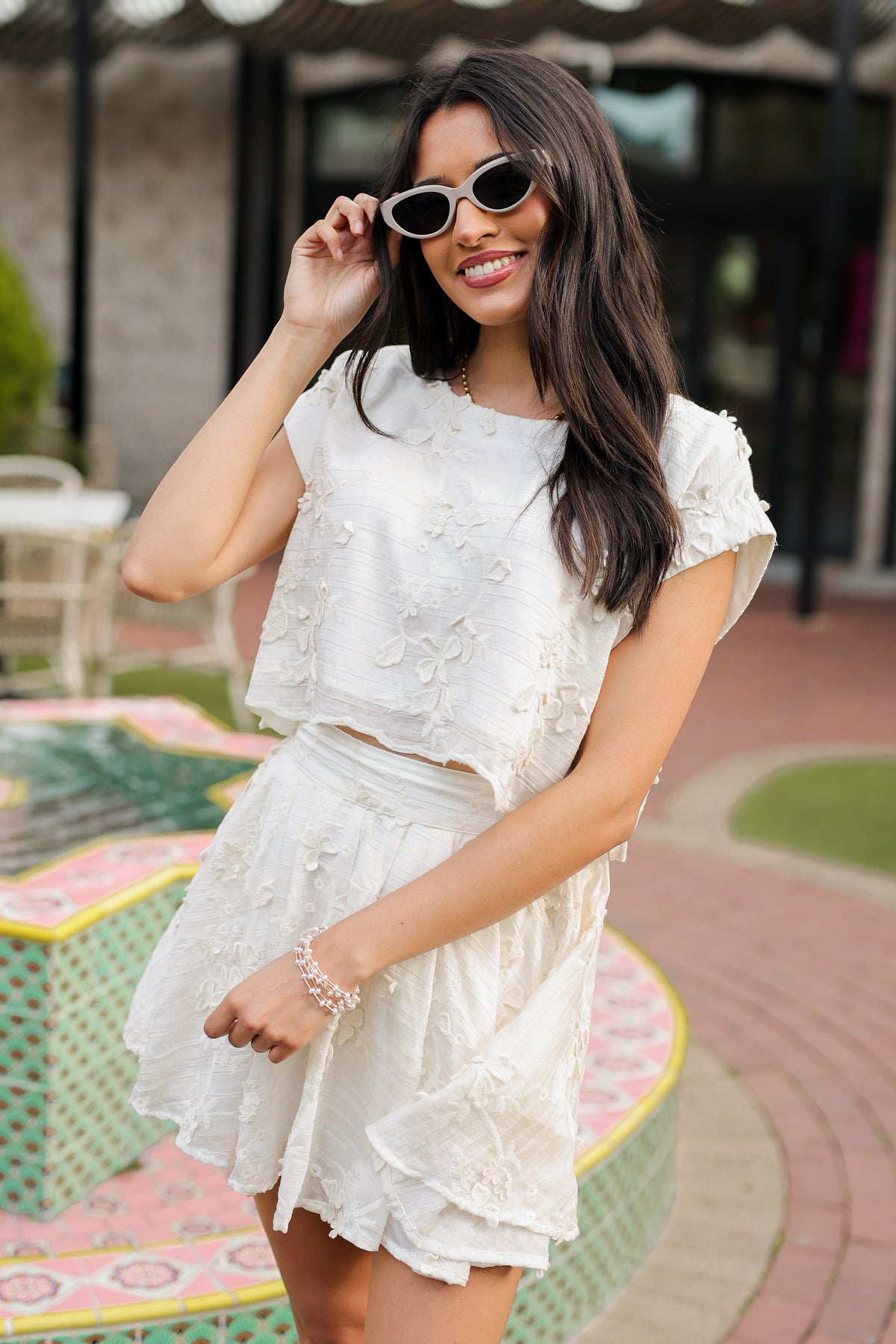 Woman in a white lace dress and sunglasses standing outdoors.