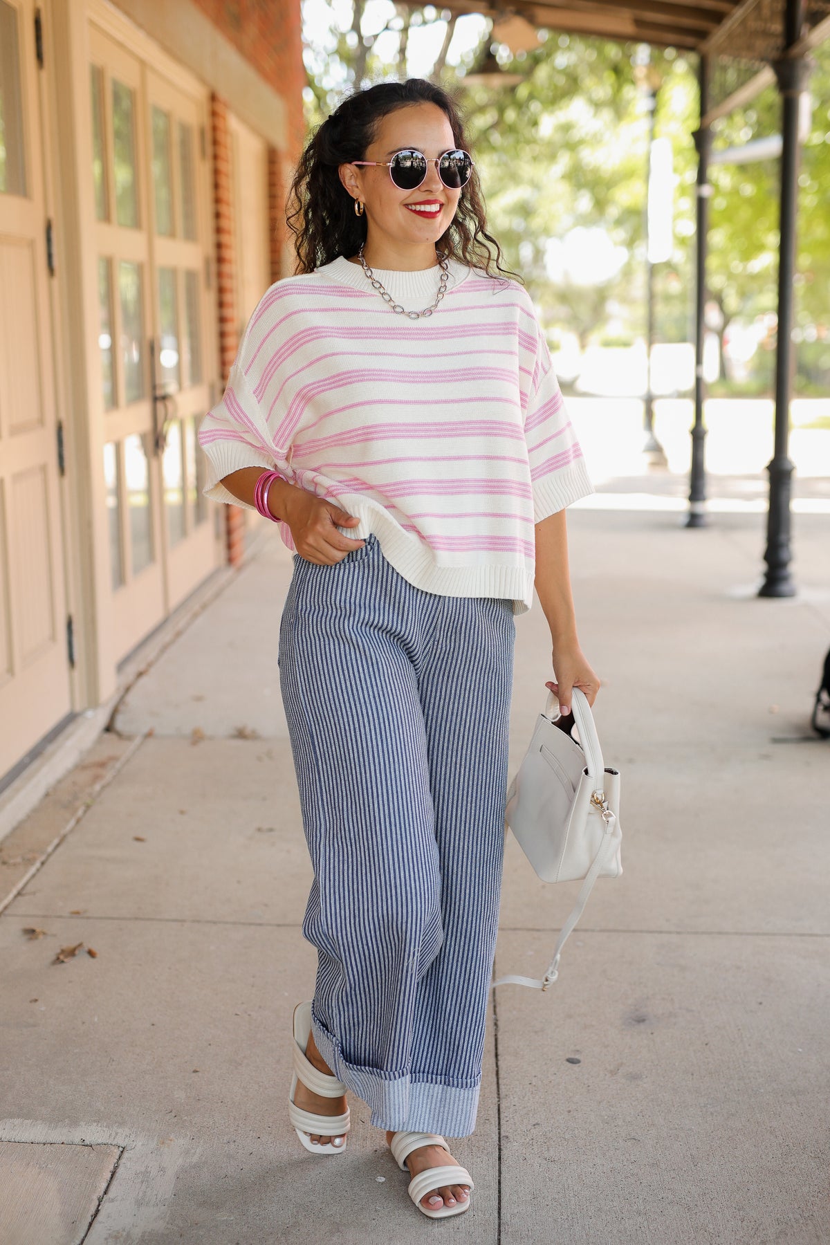 A woman walking outdoors on a sidewalk wearing a pink and white striped short-sleeve top, blue and white vertically striped wide-leg pants, white heeled sandals, and holding a white handbag.