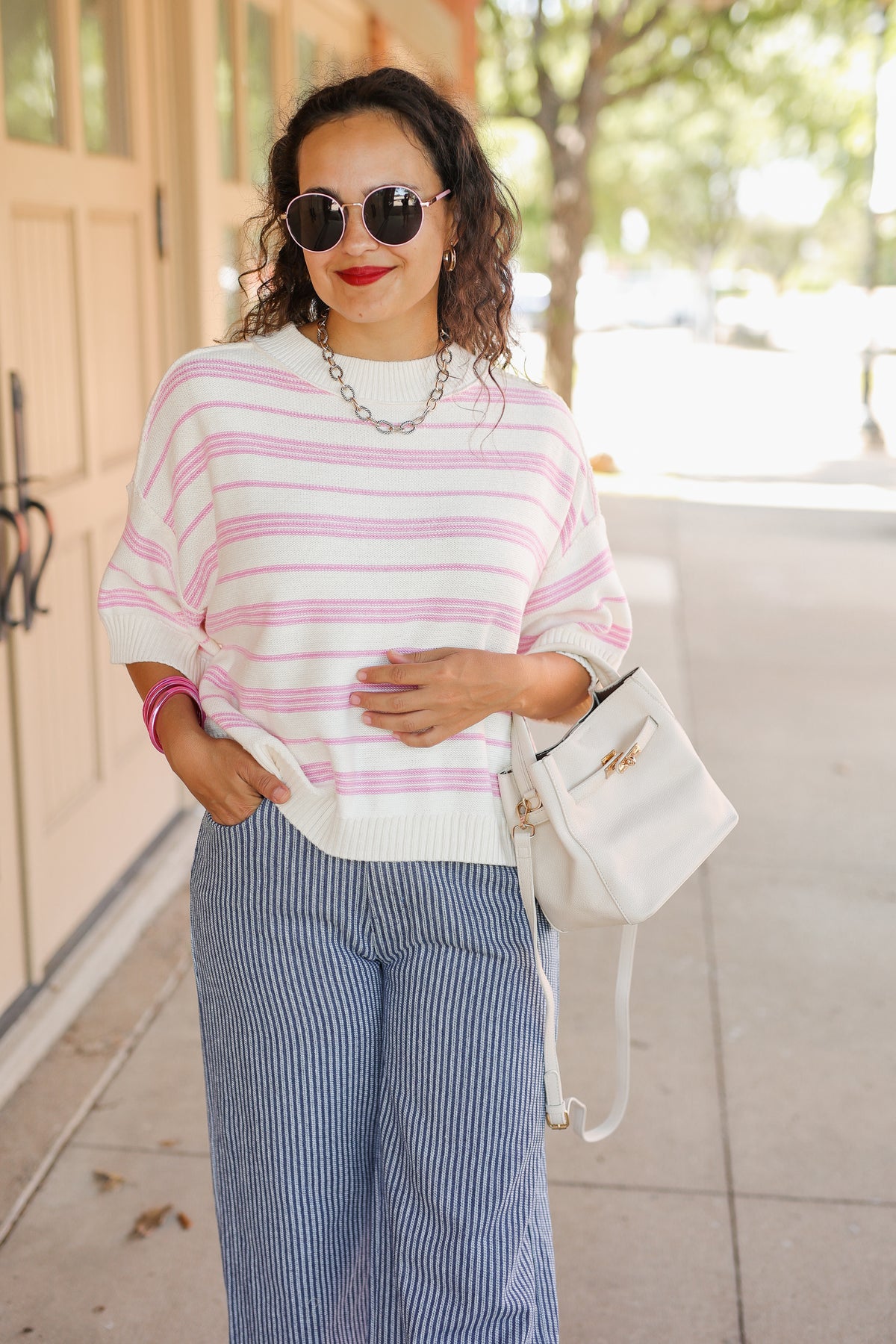 A woman wearing a white knit sweater with pink horizontal stripes, blue and white vertically striped wide-leg pants, and carrying a white shoulder bag, standing outdoors on a sidewalk.