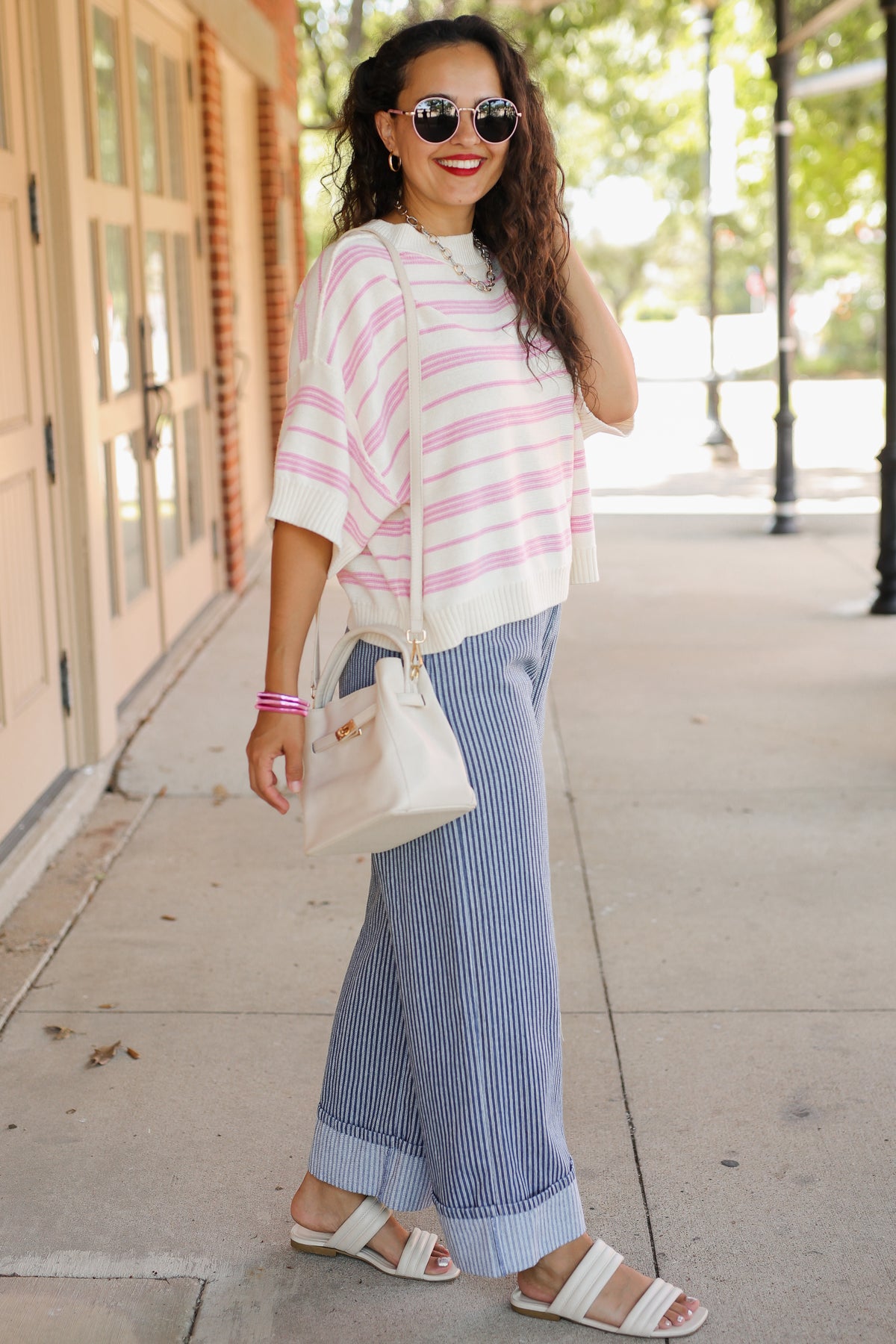 A woman stands on a city sidewalk wearing a white short-sleeve knit top with pink horizontal stripes, blue-and-white vertically striped wide-leg pants, white slide sandals, and carrying a small white shoulder bag.