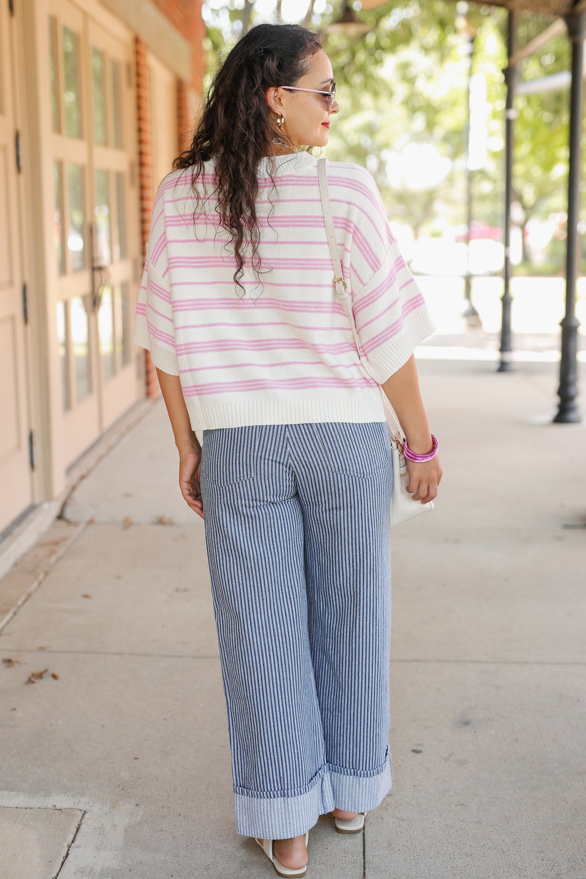 A woman is shown from behind wearing a pink and white striped knit top and blue-and-white vertically striped wide-leg pants, walking on a sunny sidewalk.