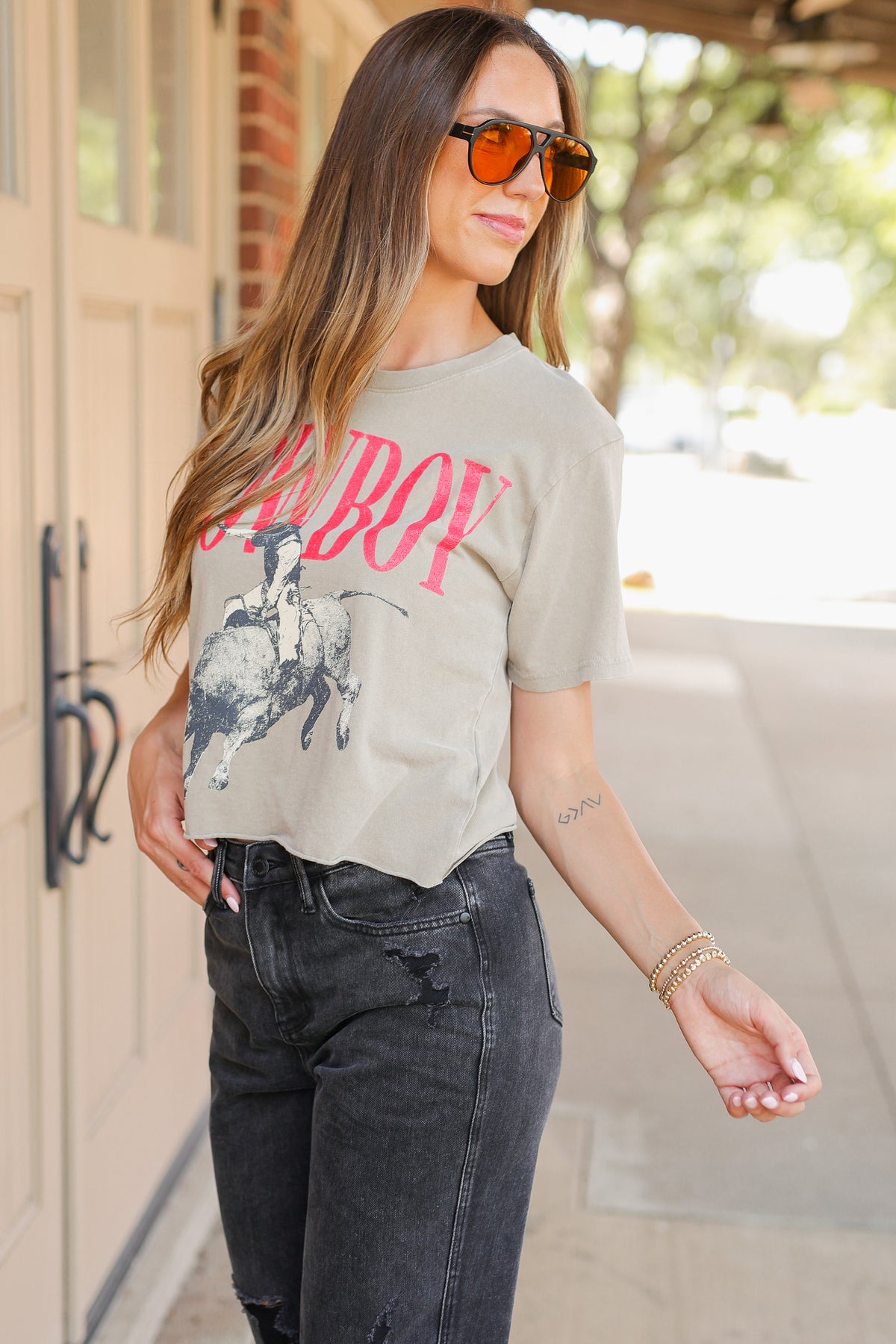 A woman wearing a light beige graphic t-shirt with a pink 'COWBOY' print and a black-and-white cowboy on a bucking horse, paired with dark gray jeans and orange-tinted sunglasses, standing outdoors.