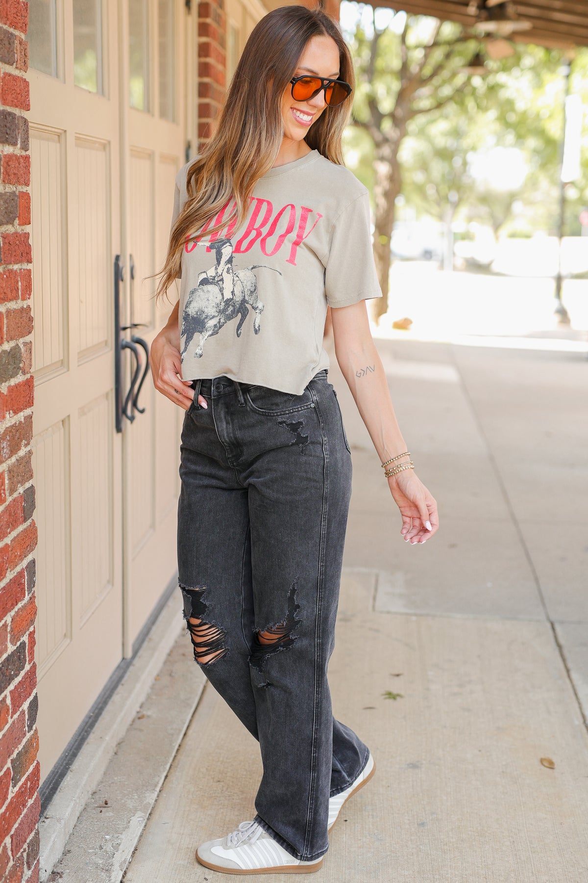 A woman wearing a light beige graphic t-shirt with a pink 'CUBO!' print, dark gray distressed jeans, and white sneakers, standing on a sidewalk next to a brick building.