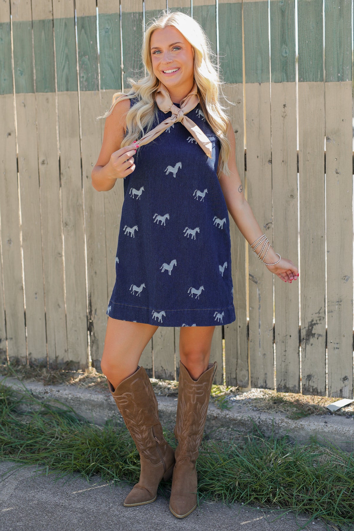 Blonde model standing outside wearing a sleeveless, denim dress with horse prints and brown cowboy boots.