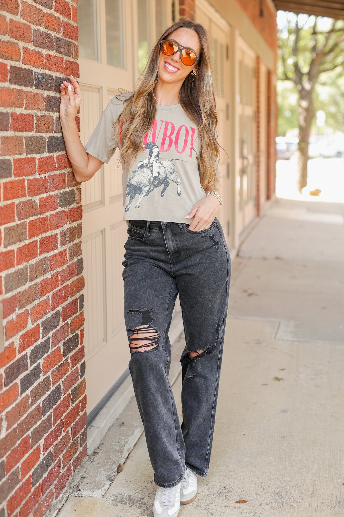 A woman stands on a sidewalk next to a red brick wall, wearing a light gray graphic t-shirt with pink text, dark gray distressed jeans, and white sneakers.