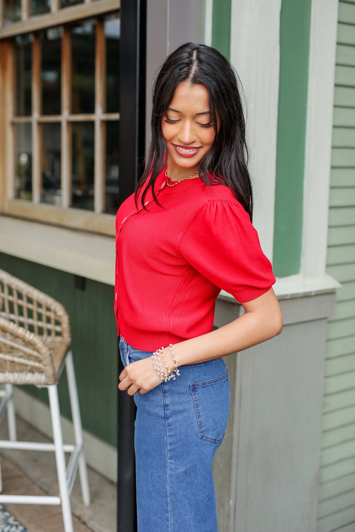 Woman in a red blouse and blue jeans standing outside a building with green walls.