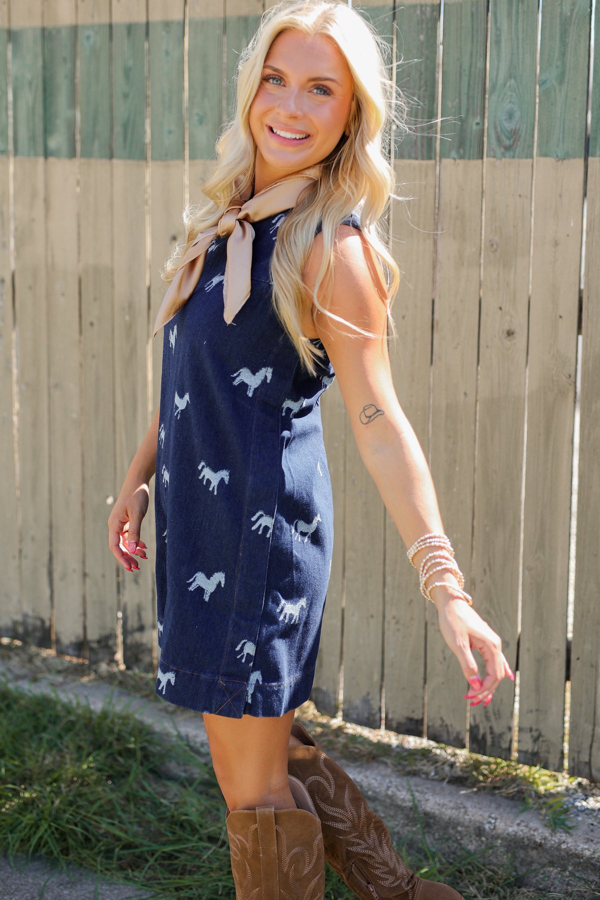 Blonde model standing outside wearing a sleeveless, denim dress with horse prints and brown cowboy boots.