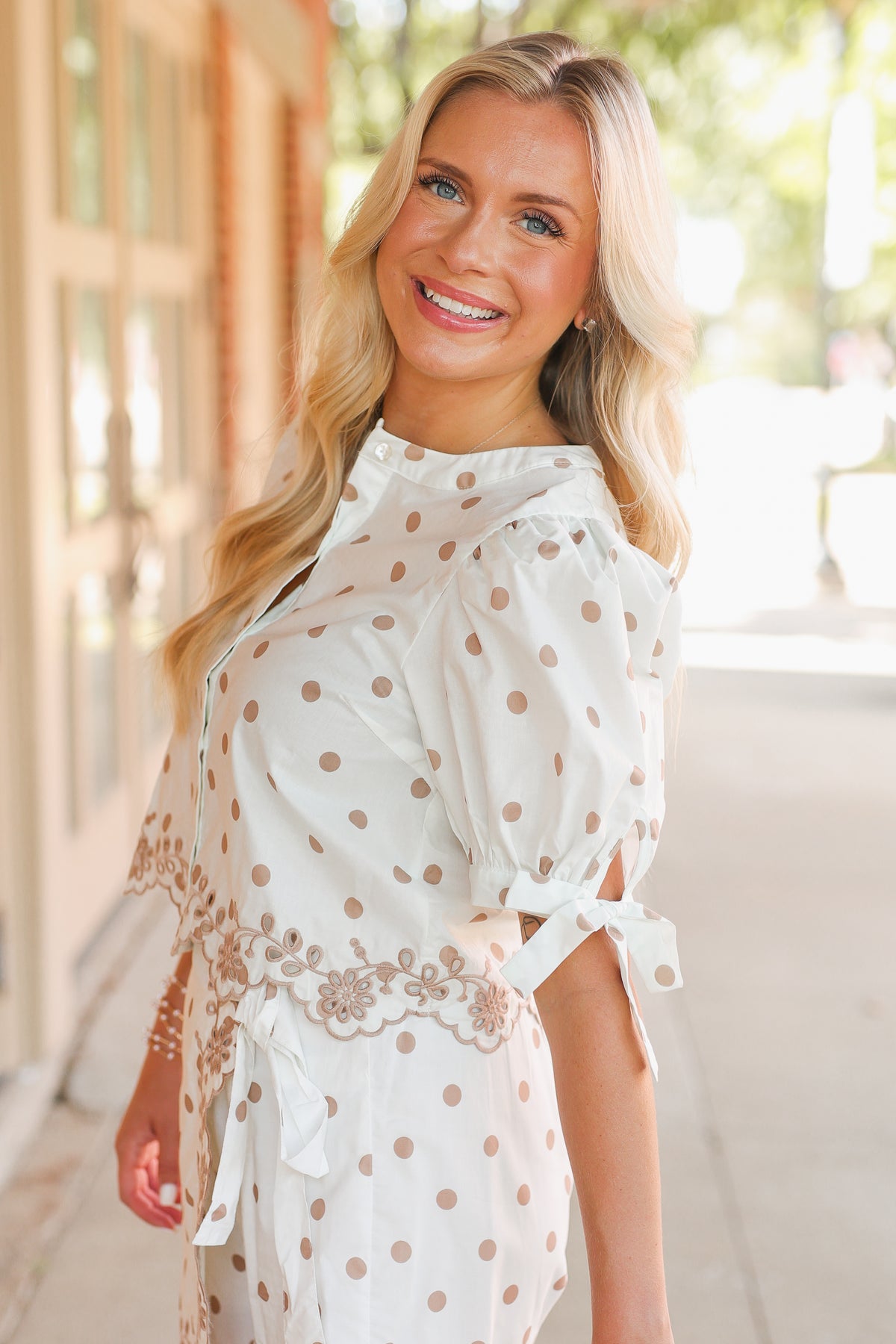 Blonde model standing outside holding a brown purse wearing a cream top and wrap skort set with taupe dots. The set also includes a scalloped hem.