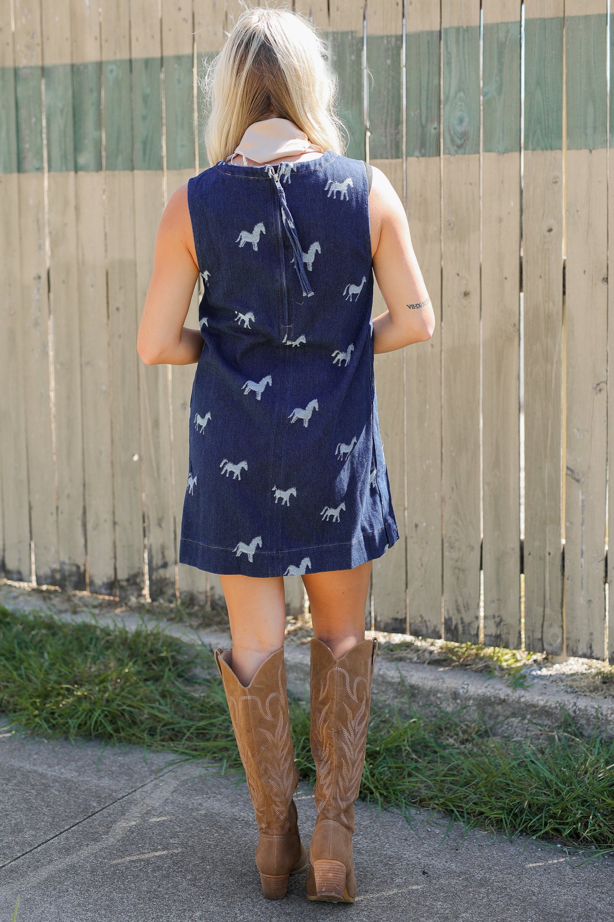 Blonde model standing outside wearing a sleeveless, denim dress with horse prints and brown cowboy boots.