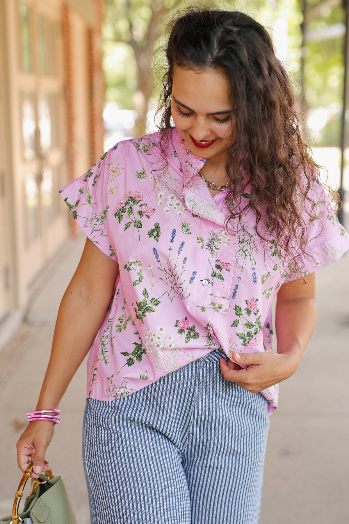 A woman wearing a light pink short-sleeve button-up blouse with a green and purple floral print, paired with blue and white vertically striped pants, walking outdoors.