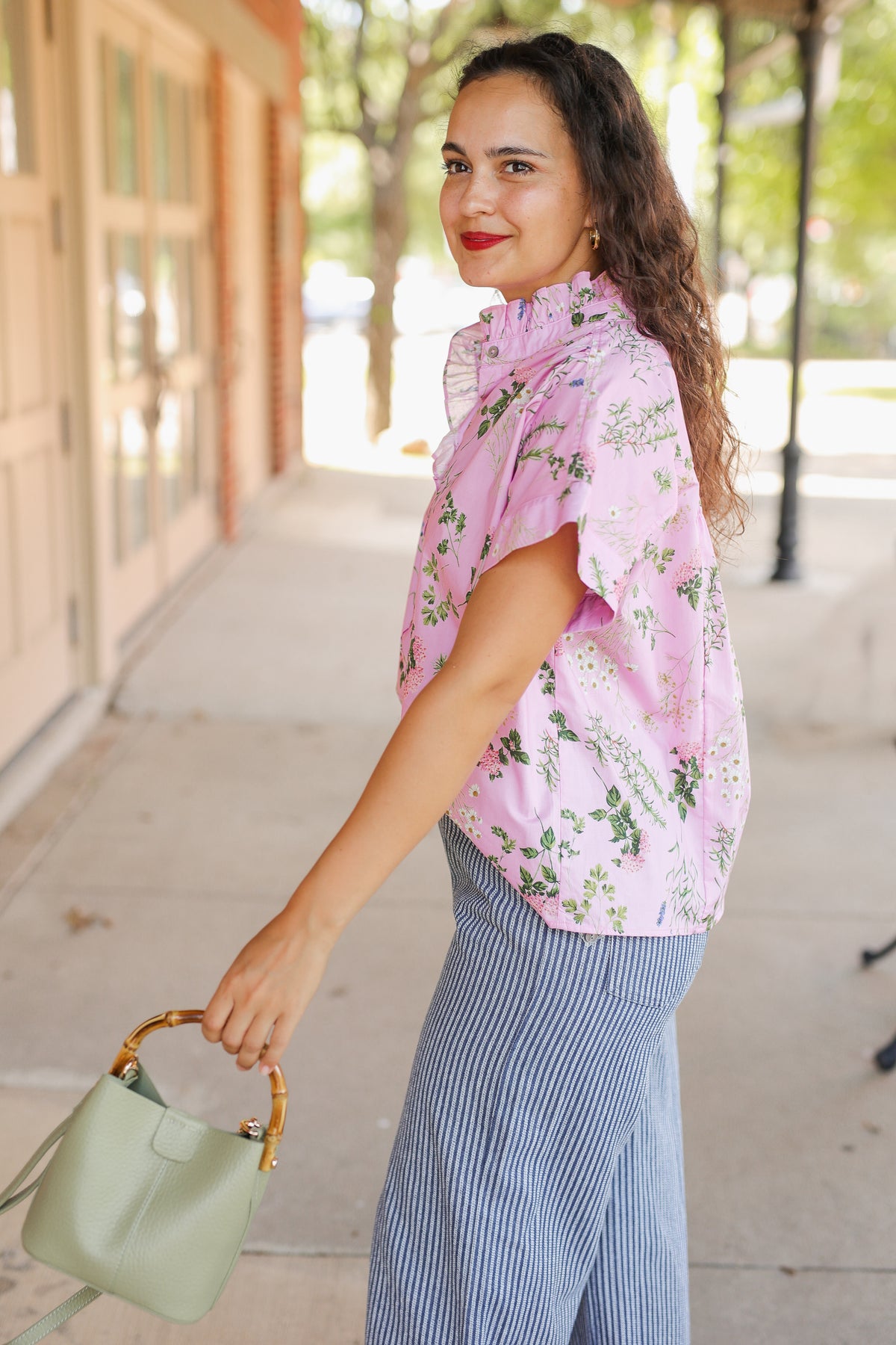 A woman wearing a pink floral short-sleeve blouse and blue-and-white striped wide-leg pants, holding a sage green handbag on a sunny sidewalk.