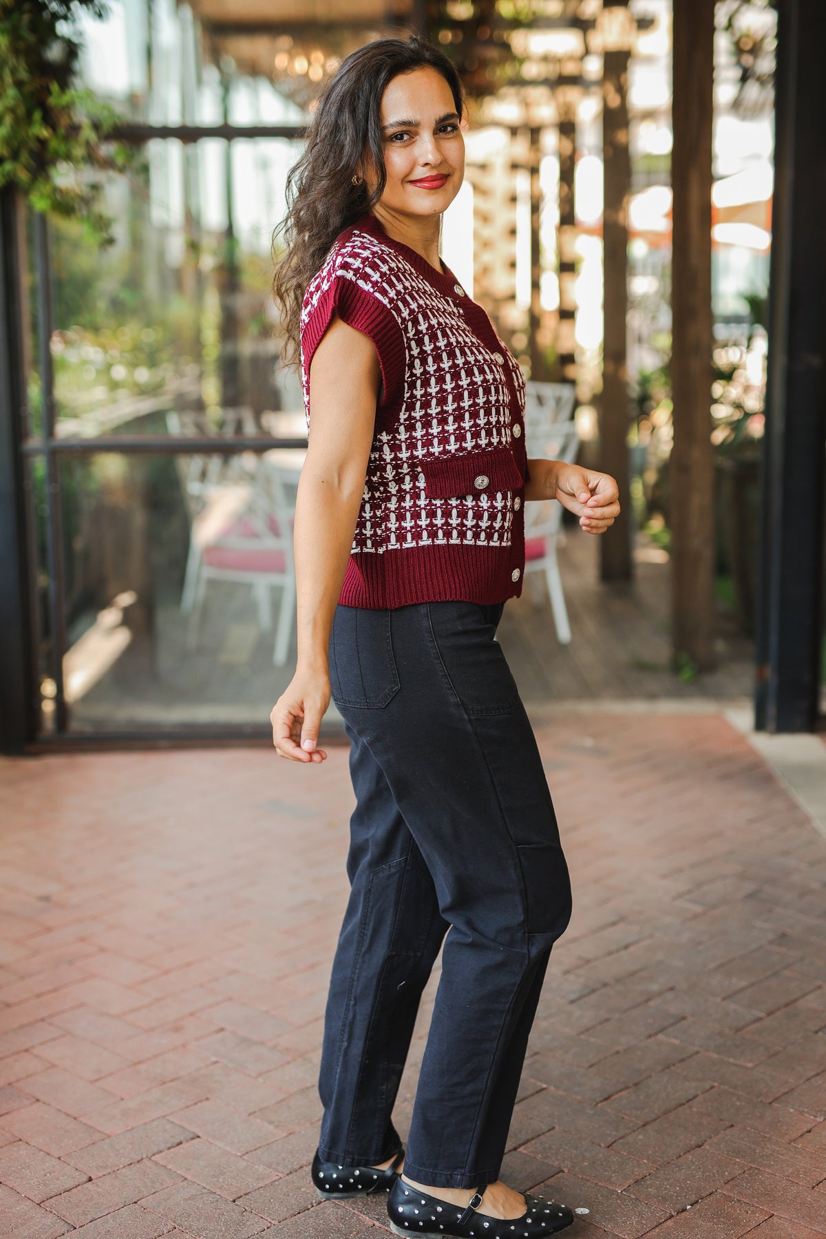 Woman wearing a patterned top and dark pants standing on a brick pavement.