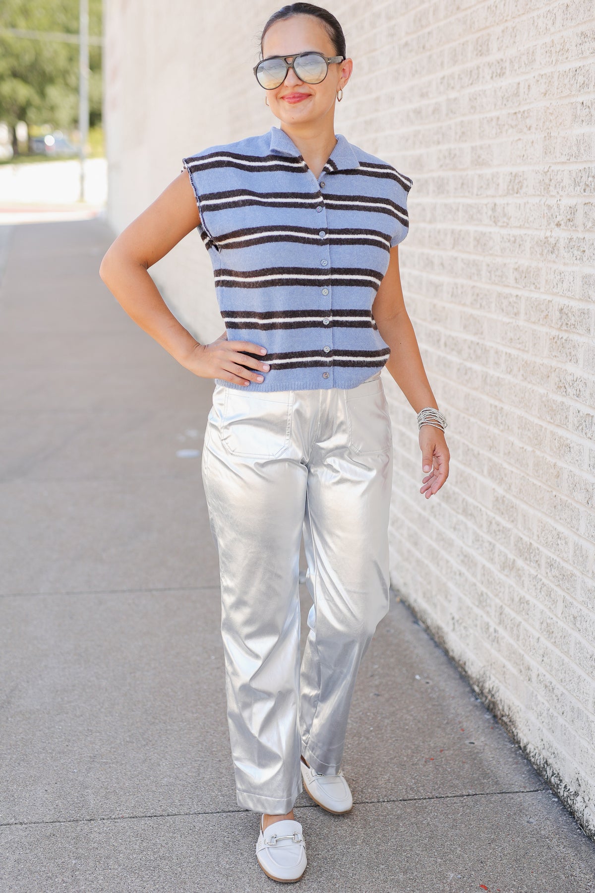 Woman standing outside wearing a blue striped sweater vest and metallic silver pants.