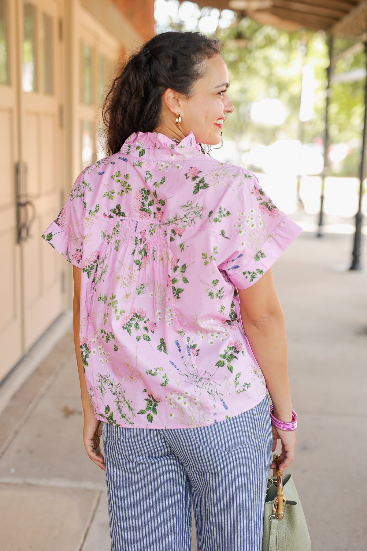 A woman wearing a light pink short-sleeve blouse with a green and white floral print, paired with blue and white vertically striped pants, standing outdoors on a sunny day.