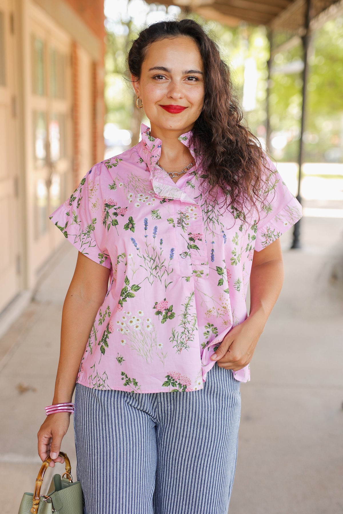 A woman wearing a light pink short-sleeve button-up blouse with a green and white botanical floral print, paired with blue and white vertically striped pants, standing on a sunny outdoor porch.