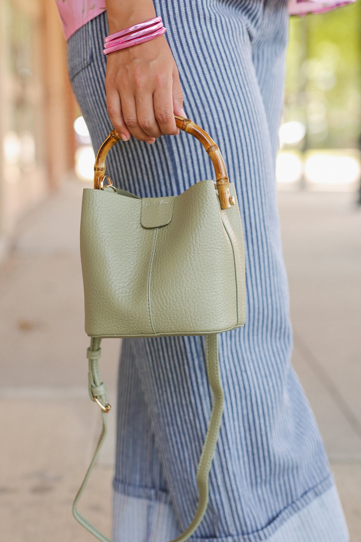 A person outdoors holding a small pebbled leather bucket bag with a light sage green body and natural bamboo top handle. The person is wearing blue and white striped pants and pink bracelets.