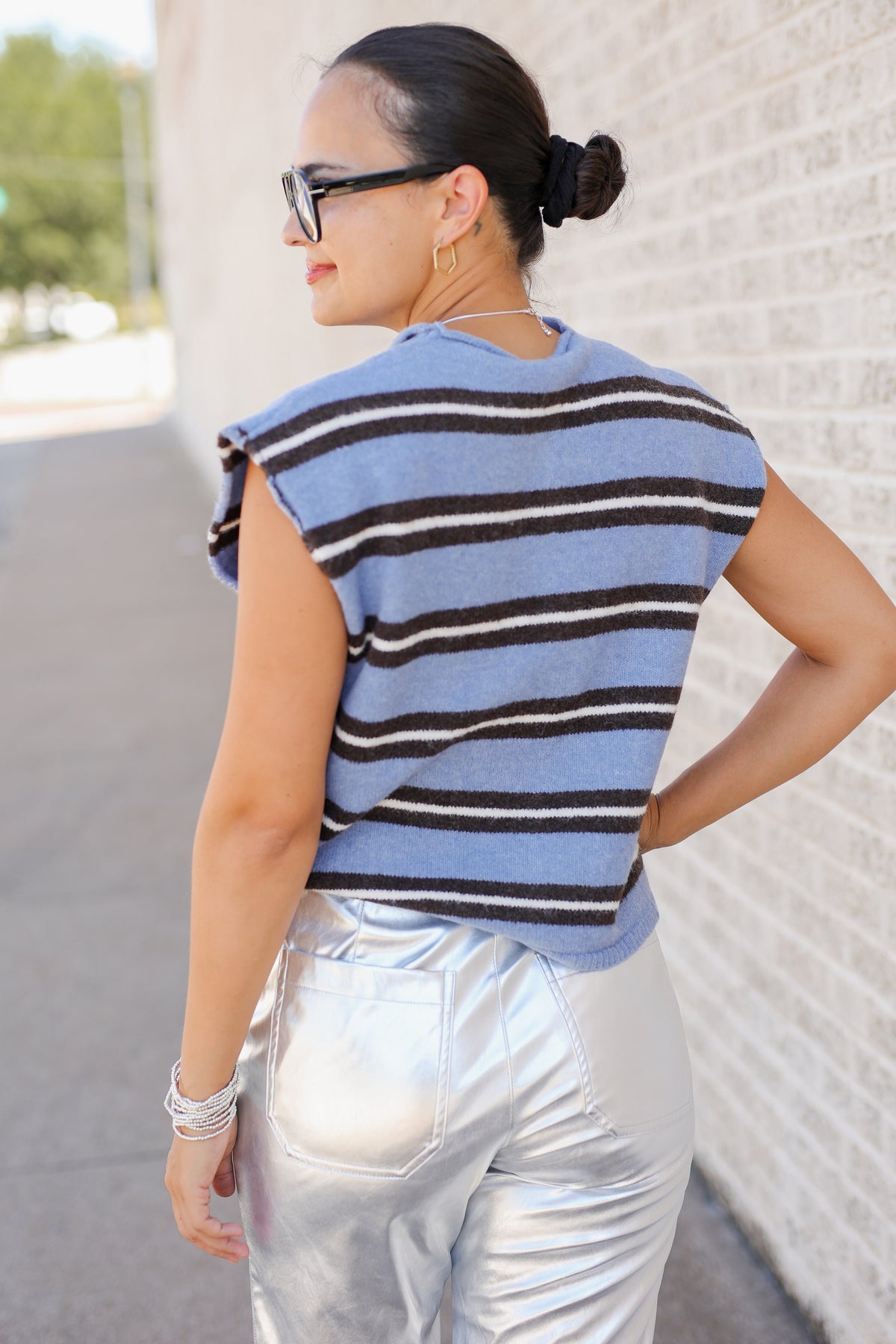 A woman wearing a light blue sleeveless knit top with dark brown and white horizontal stripes, paired with shiny silver pants, photographed outdoors against a light brick wall.