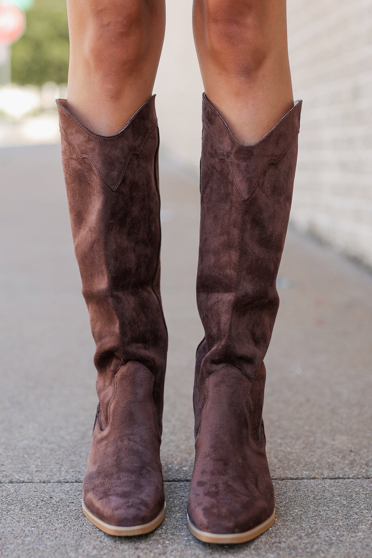 A person wearing knee-high brown suede boots with a western-style pointed toe and low block heel, standing on a paved sidewalk.