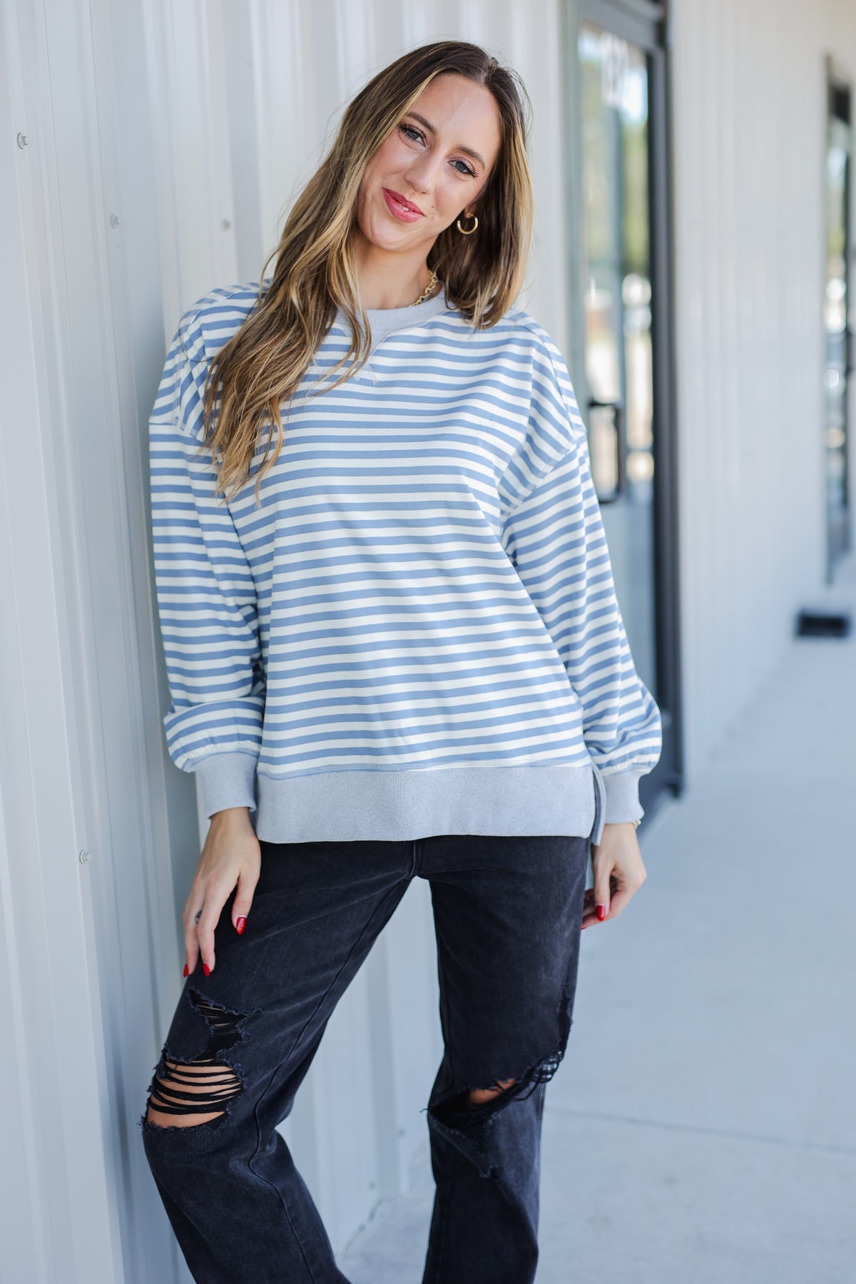 Woman wearing a striped sweater and black pants leaning against a white wall.