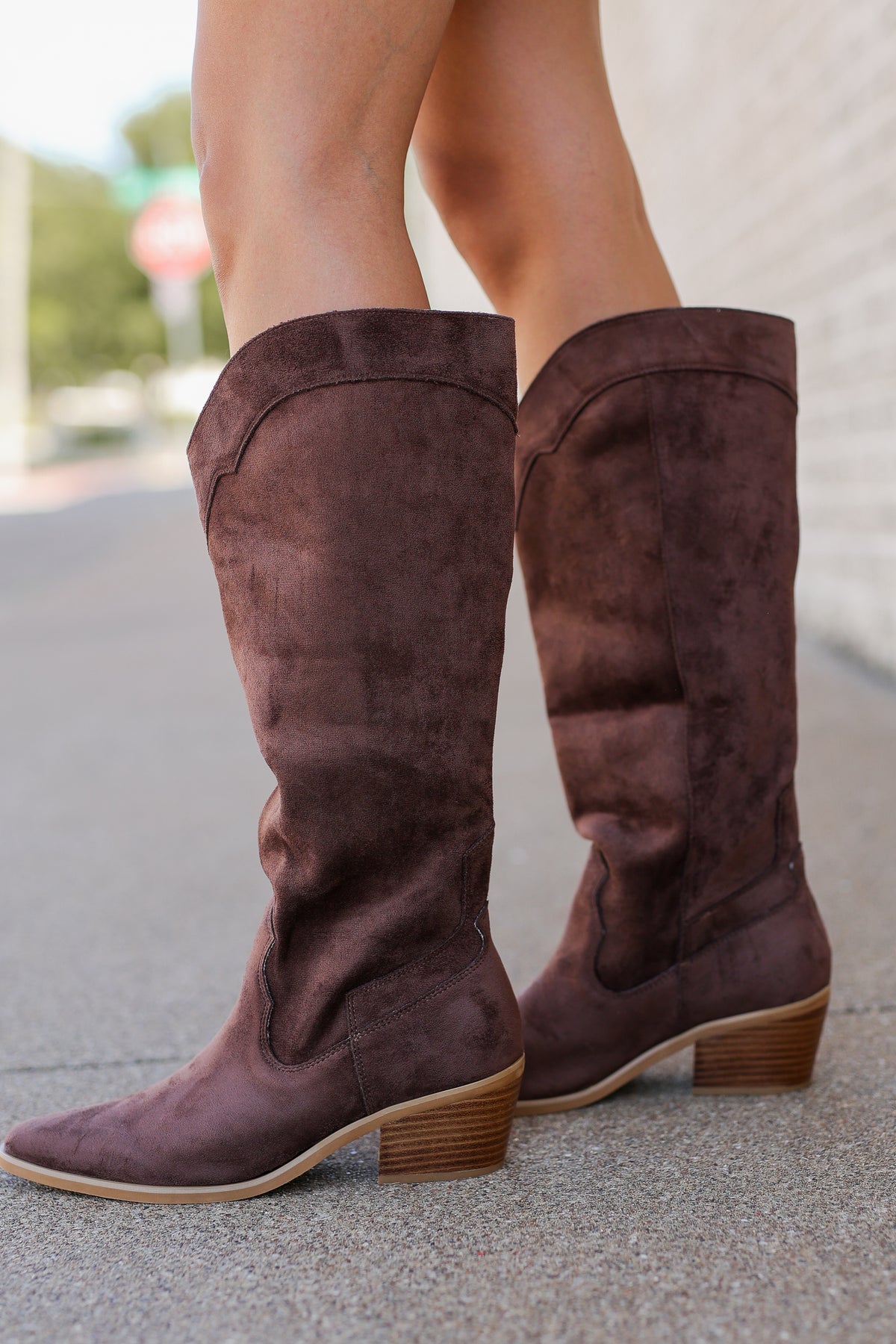 A pair of knee-high brown suede boots with low stacked block heels, shown on a model standing on a paved outdoor walkway.