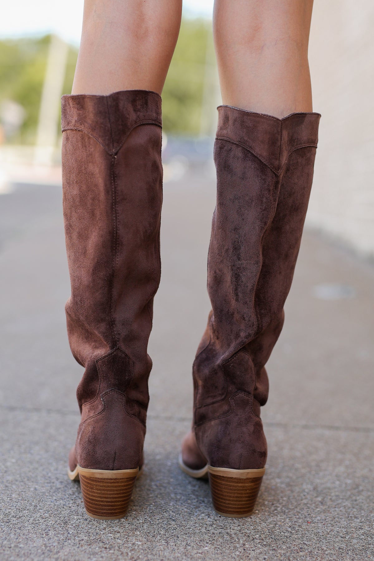A pair of knee-high brown suede boots with stacked wooden block heels, shown from the back on a person standing on a paved outdoor walkway.