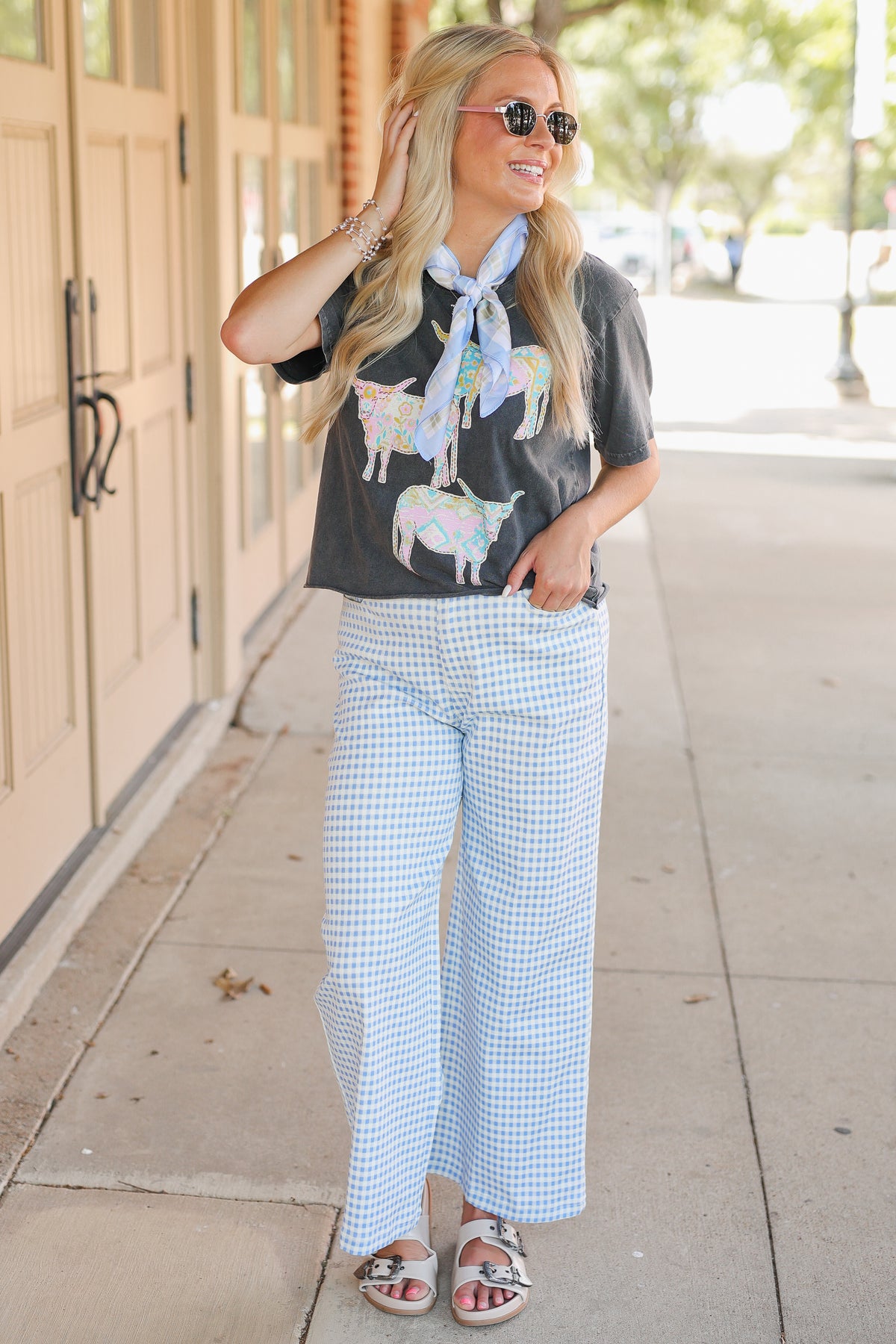 Blonde model wearing a black graphic tee, a blue scarf, and blue gingham pants, standing outside.