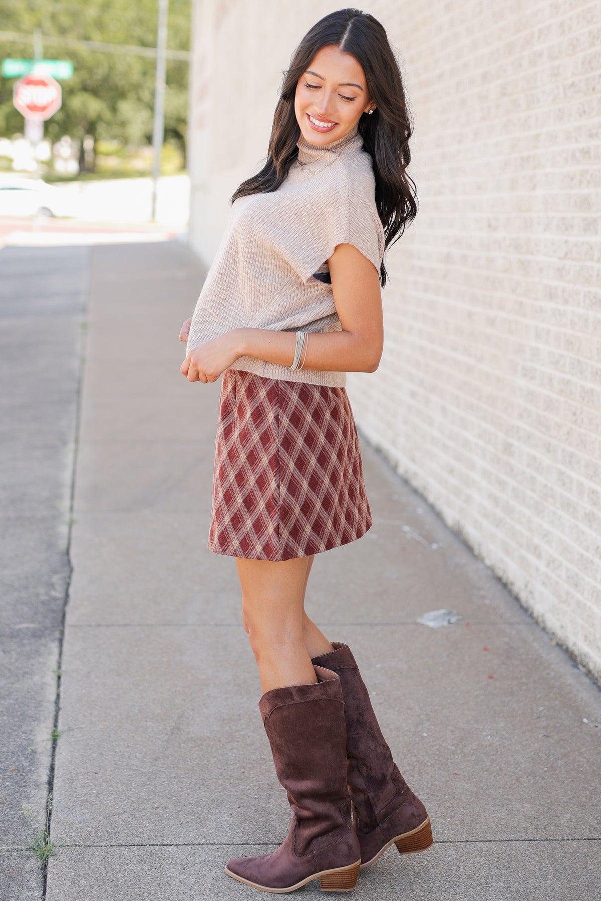 A woman posing on a sidewalk next to a white brick wall, wearing a beige knit mock-neck top, a rust and brown argyle skirt, and dark brown suede knee-high boots.