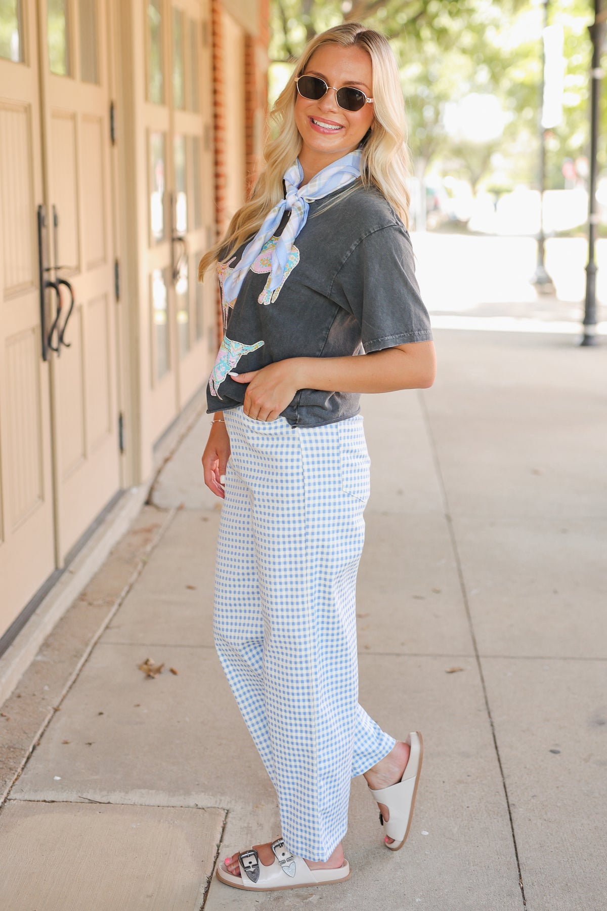 Blonde model wearing a black graphic tee, a blue scarf, and blue gingham pants, standing outside.