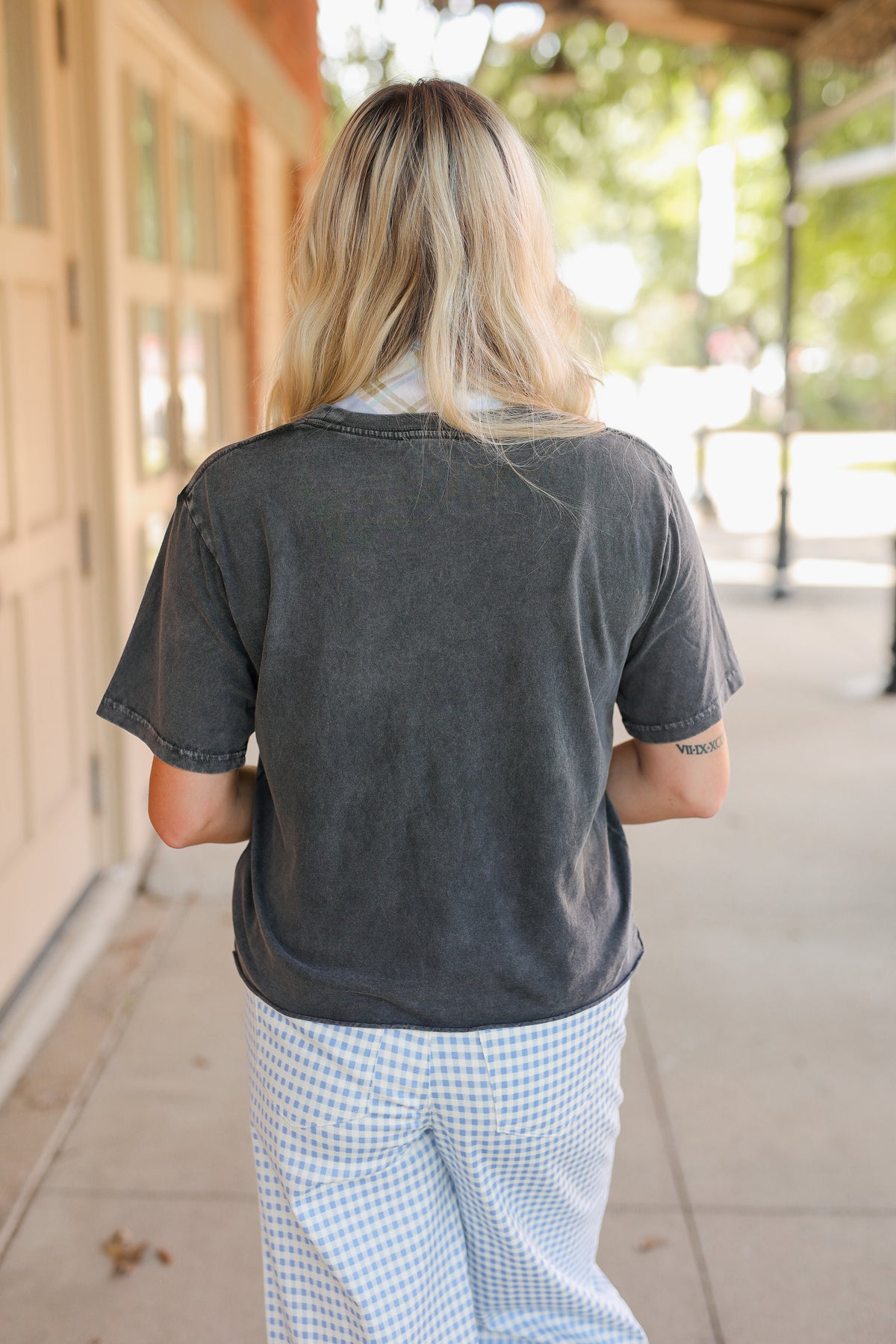 Blonde model wearing a black graphic tee, a blue scarf, and blue gingham pants, standing outside.
