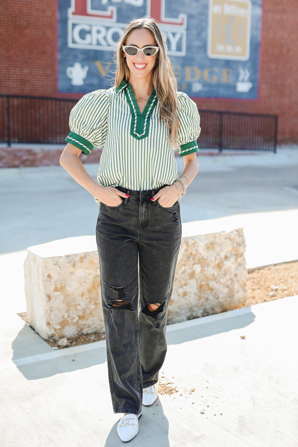 Woman wearing a striped shirt and jeans standing in front of a brick wall with a sign.